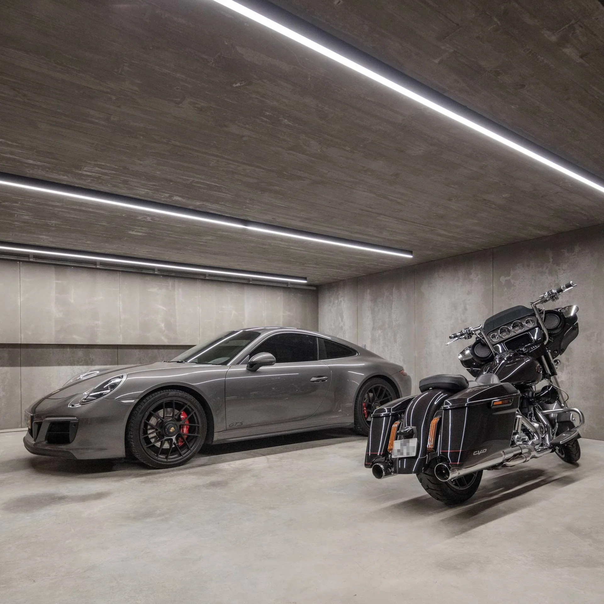 A minimalist concrete garage features a grey sports car and a black motorcycle under recessed linear ceiling lights.