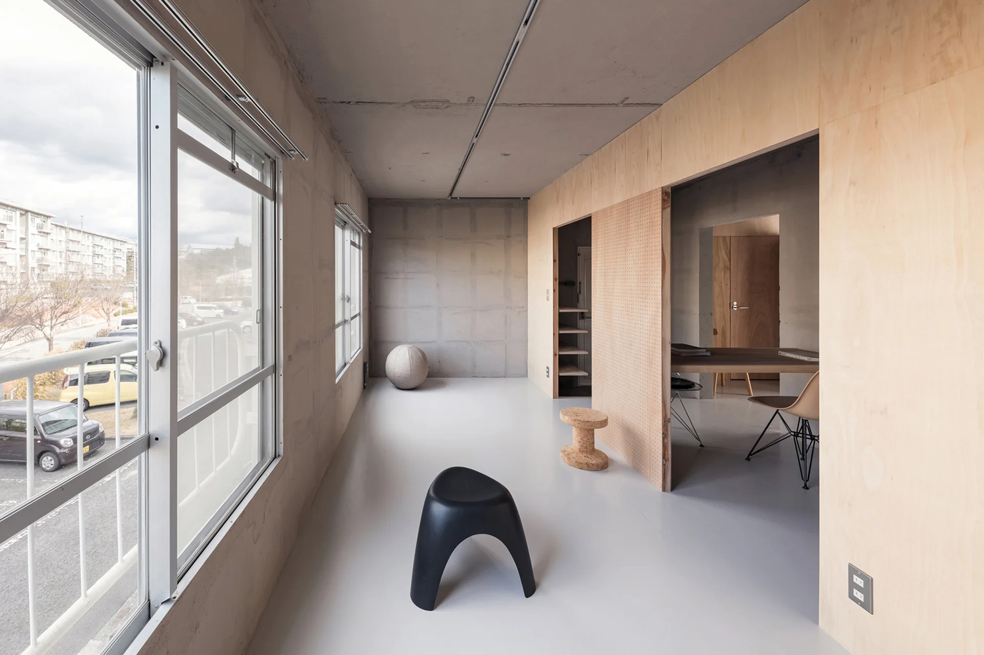 A minimalist room with raw concrete walls, a smooth gray floor, and a warm plywood partition with a sliding perforated pegboard door.