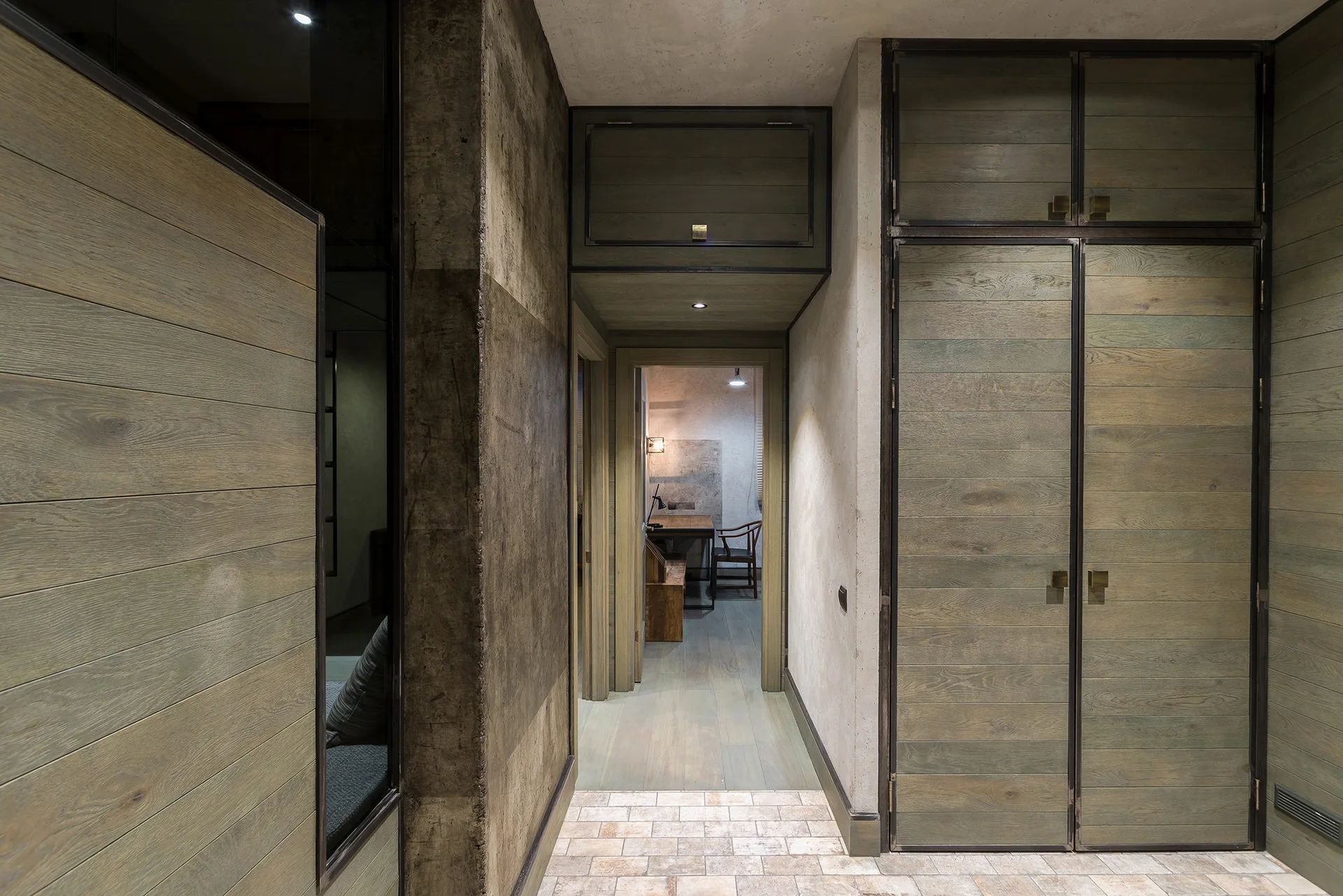 An industrial corridor features built-in cabinets with muted green wood paneling, a concrete accent wall, and stone floor tiles.