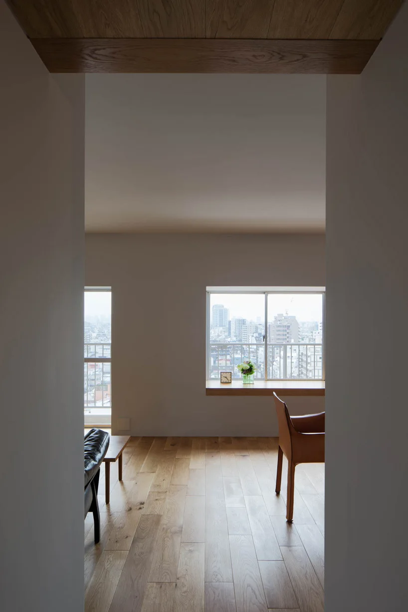 A minimalist living space framed by a doorway, featuring light wood floors, a tan leather chair, and a deep wood window sill.