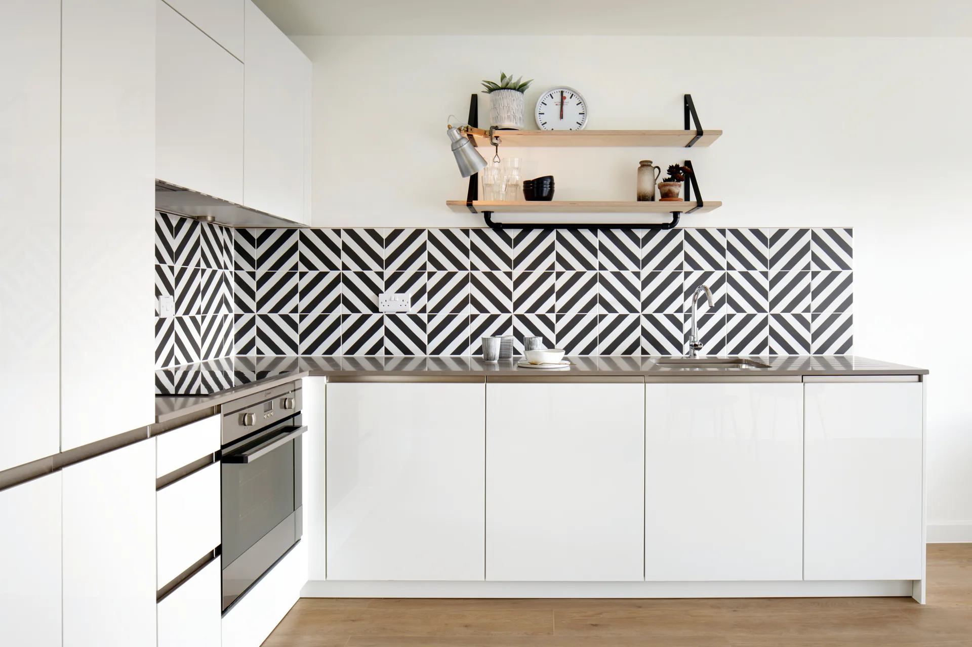 A modern kitchen featuring glossy white flat-panel cabinets, a black and white geometric tiled backsplash, and light wood floating shelves.