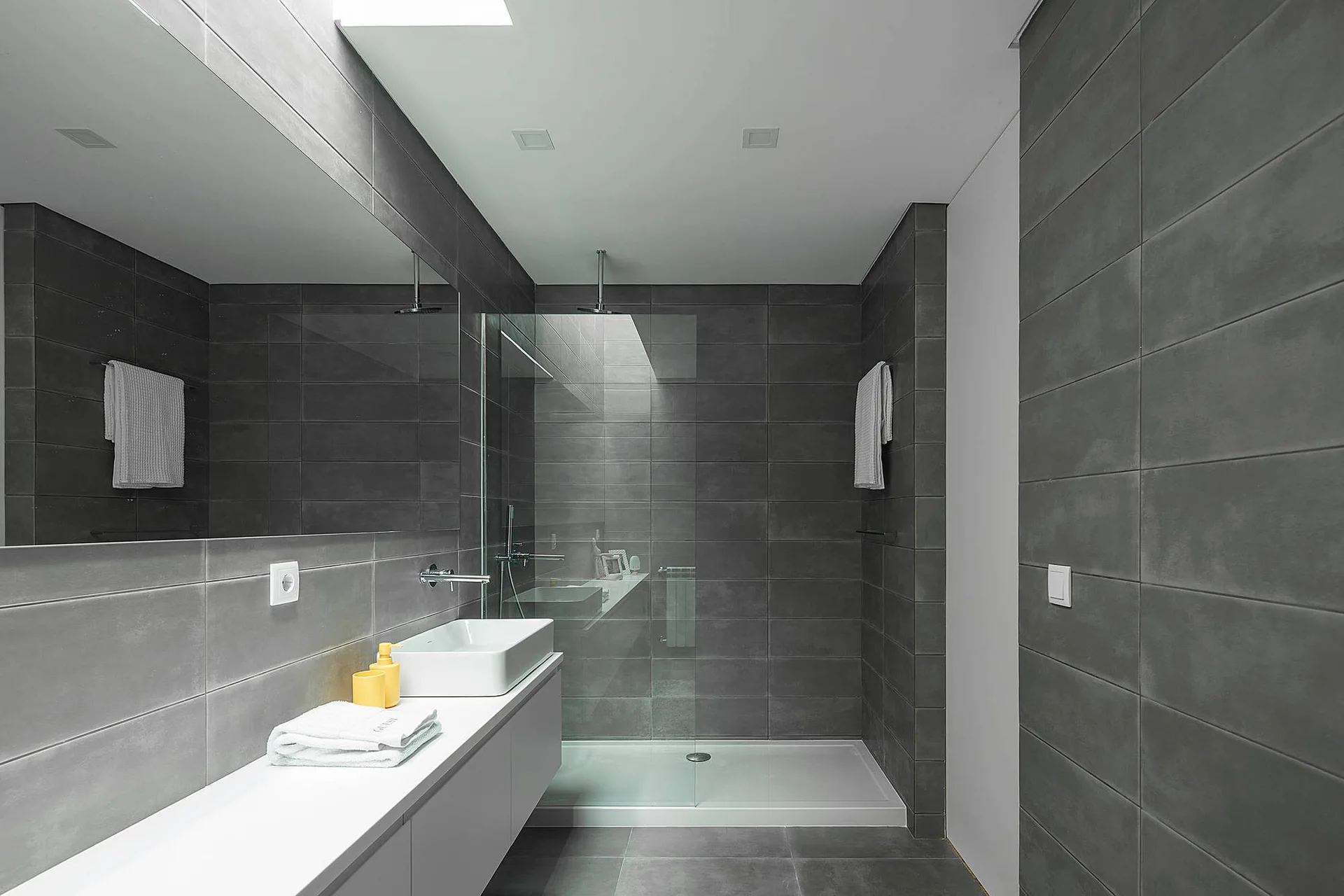 A modern bathroom with dark grey wall tiles, a white floating vanity, a vessel sink, and a glass-enclosed shower lit by a skylight.