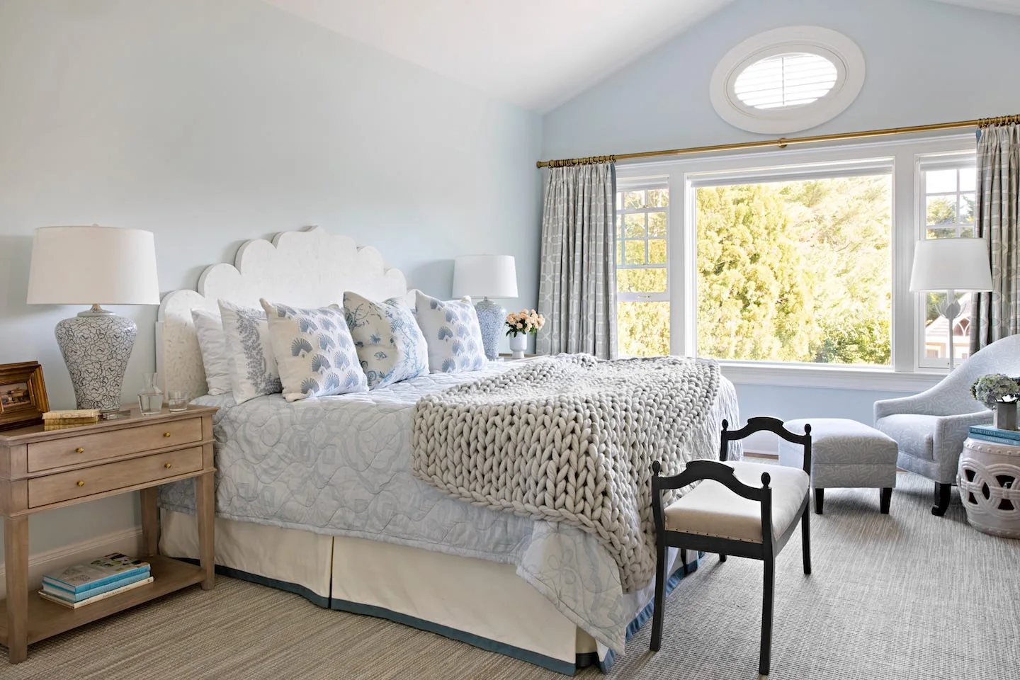 A light blue bedroom with a large bed featuring a white scalloped headboard, patterned pillows, a chunky knit throw blanket, and a large window.