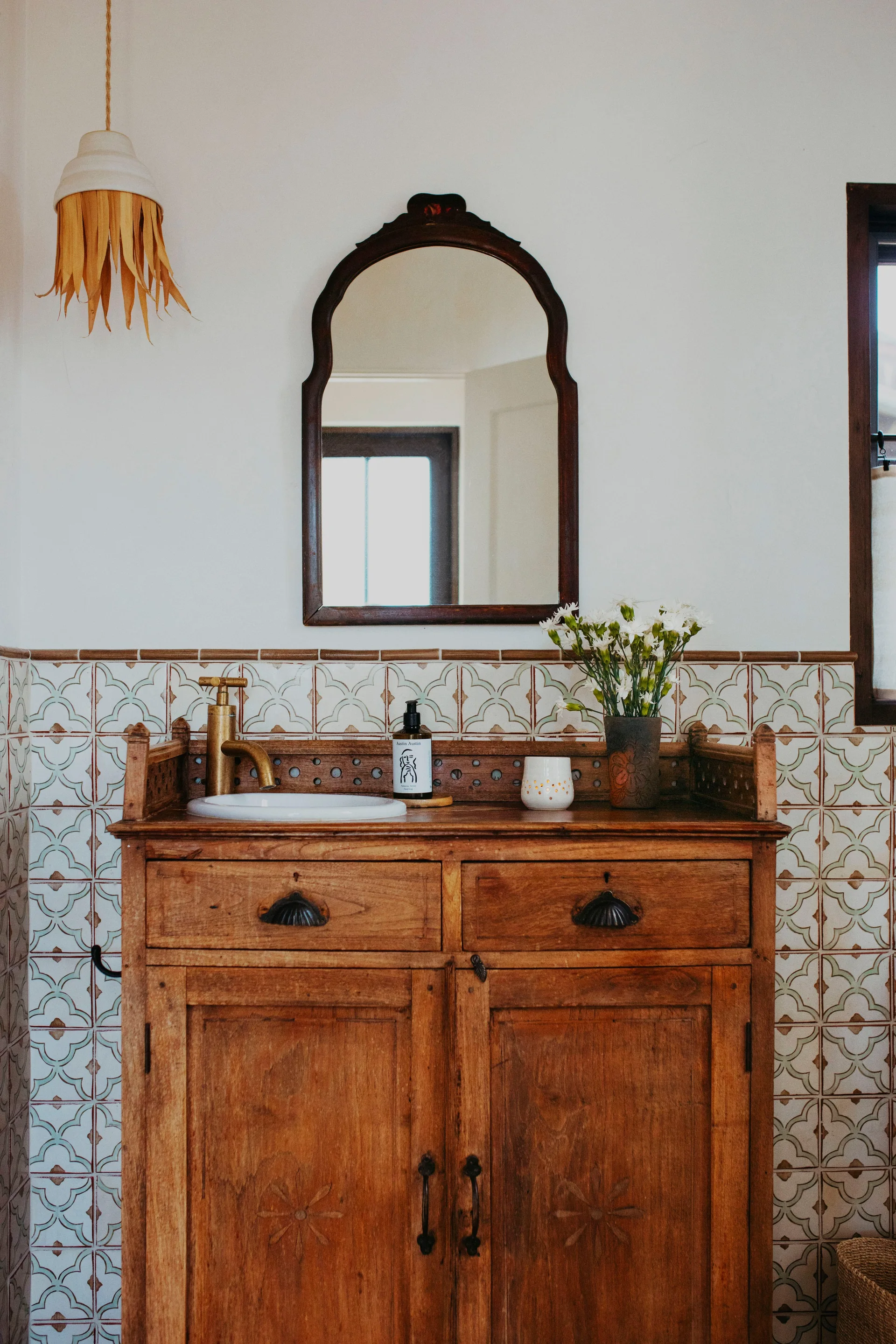 A rustic powder room features a vintage wooden washstand vanity with a brass faucet set against a patterned ceramic tile wall below an arched wood mirror.
