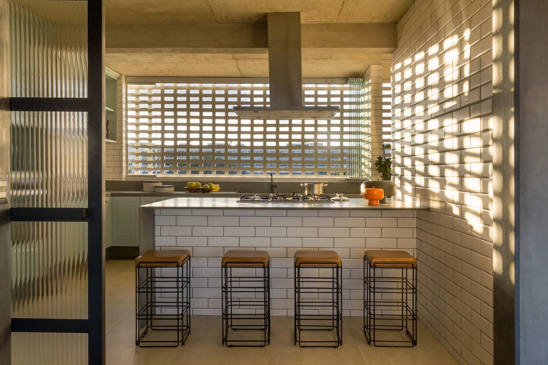 A modern industrial kitchen featuring a subway tiled island with four leather-topped bar stools and a patterned breezeblock window wall.