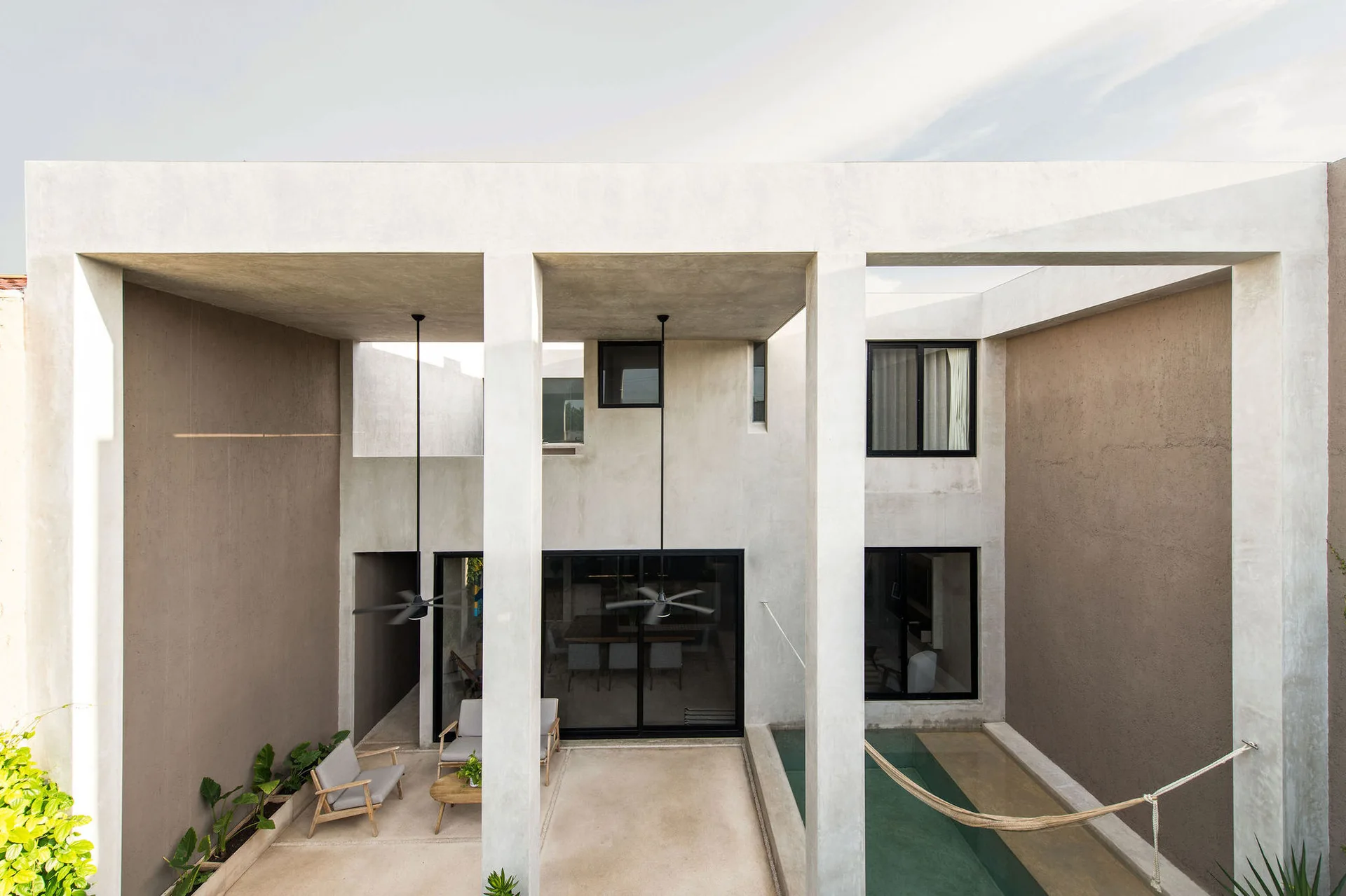 A modern concrete patio features thick columns, a narrow swimming pool with a hammock, and a small sitting area with grey armchairs.
