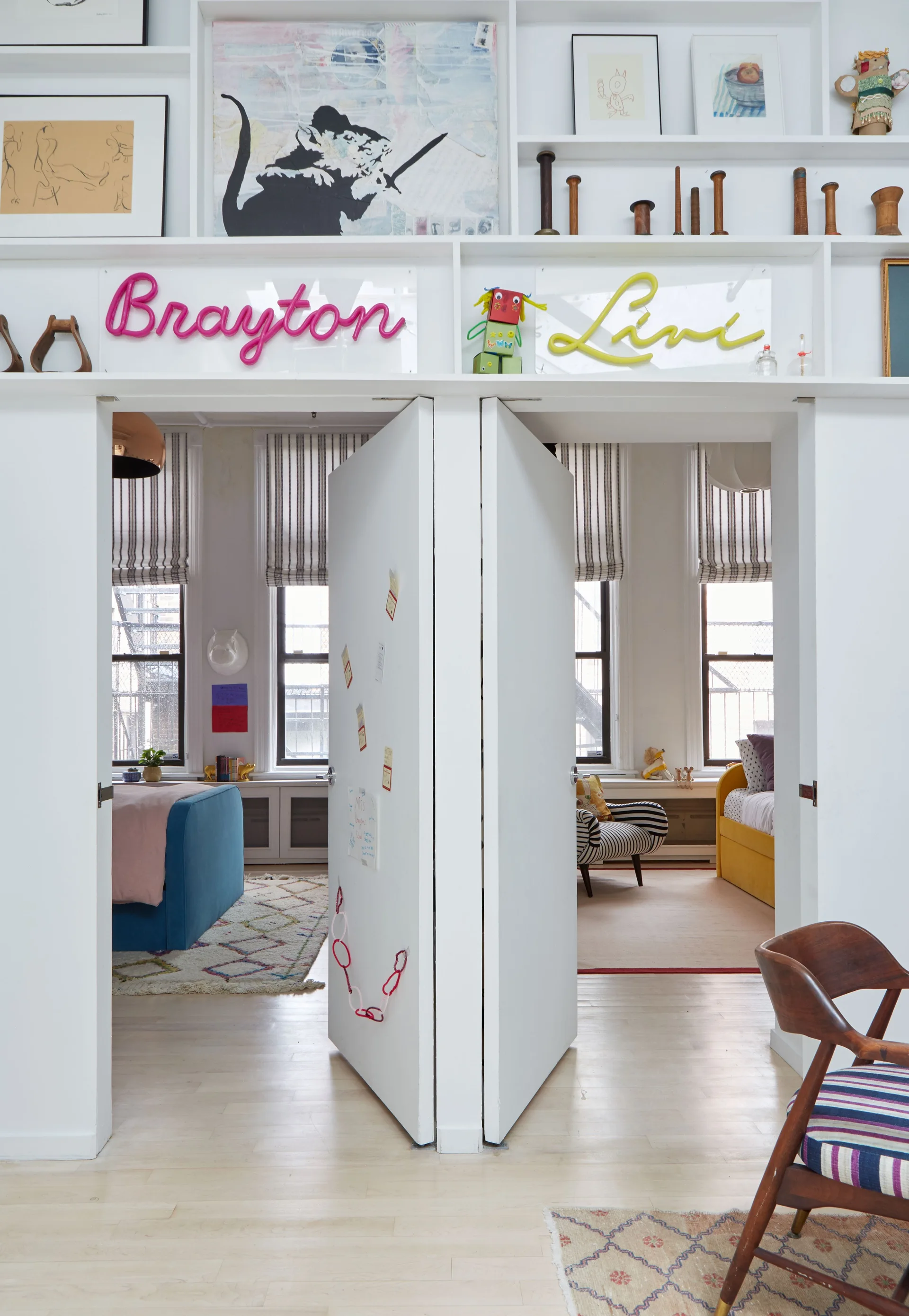 Two connected kids' bedrooms feature open white doors, personalized neon name signs, and high built-in shelving displaying artwork.