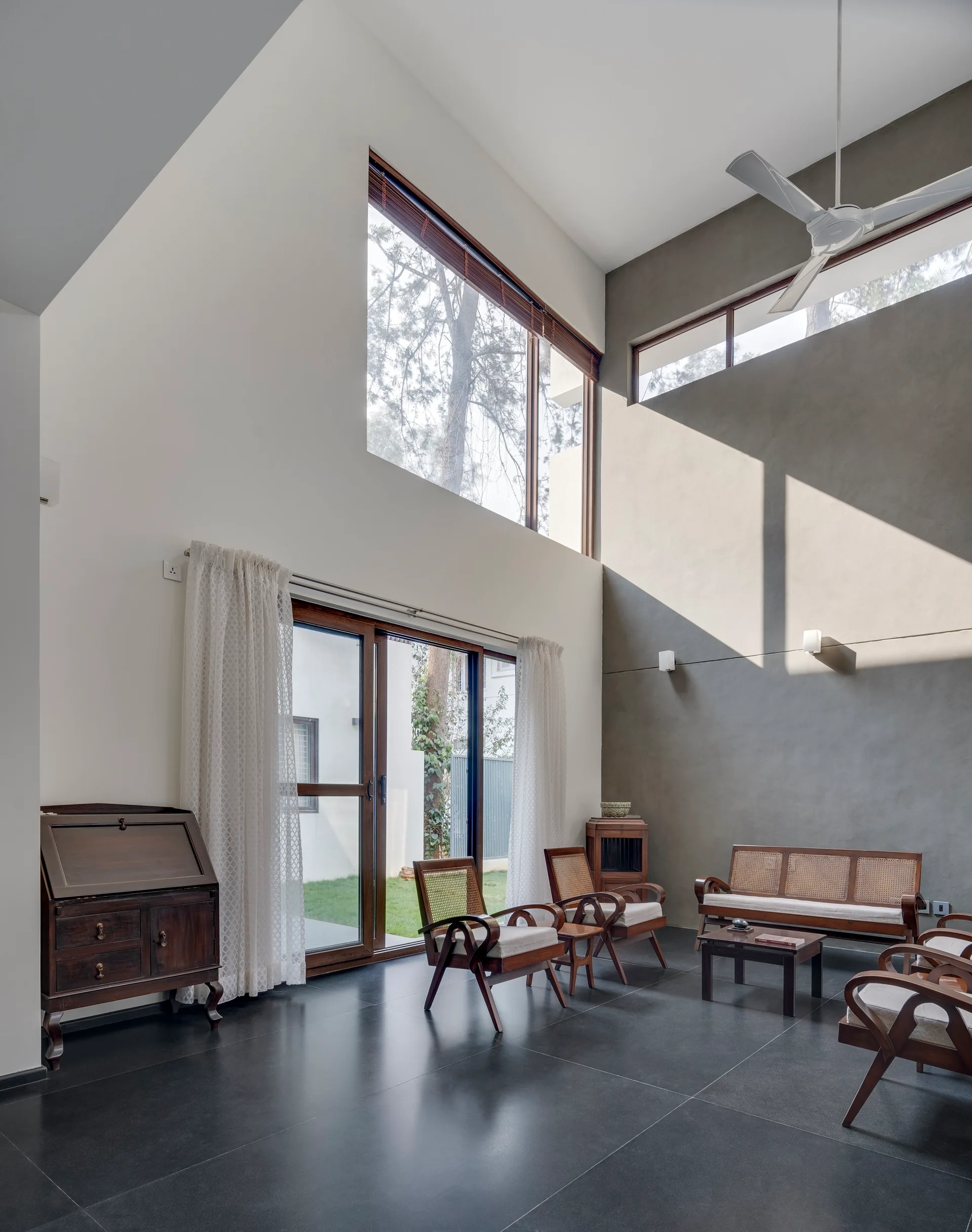 A high-ceilinged living room features dark tile flooring, mid-century wooden armchairs with cane backs, and large windows bringing in natural light.