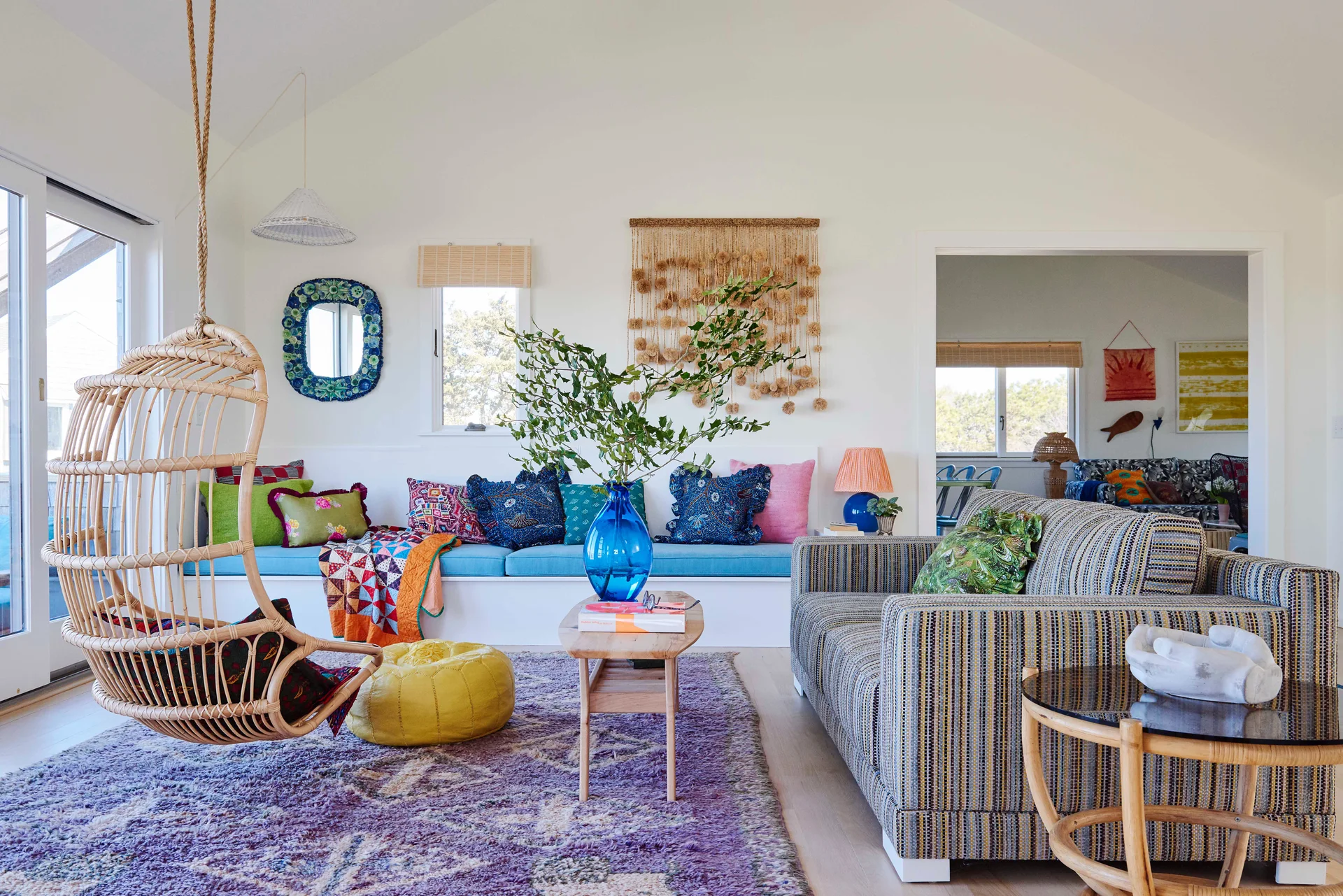 A bright living room features a hanging rattan swing, a striped sofa, a built-in window bench with colorful pillows, and a purple area rug.