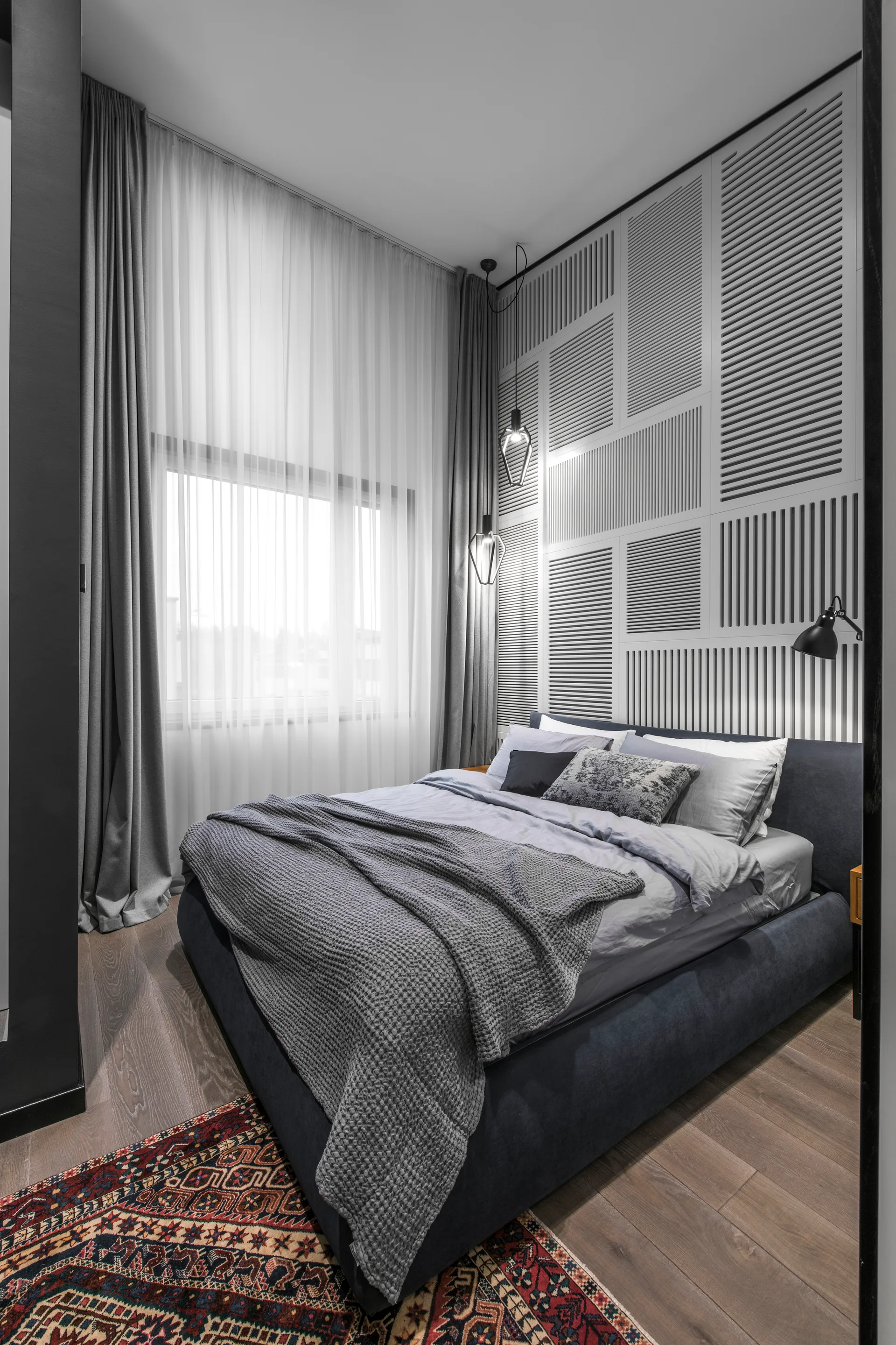 A contemporary bedroom featuring a charcoal upholstered bed, a white geometric slatted accent wall, and long grey curtains by a window.