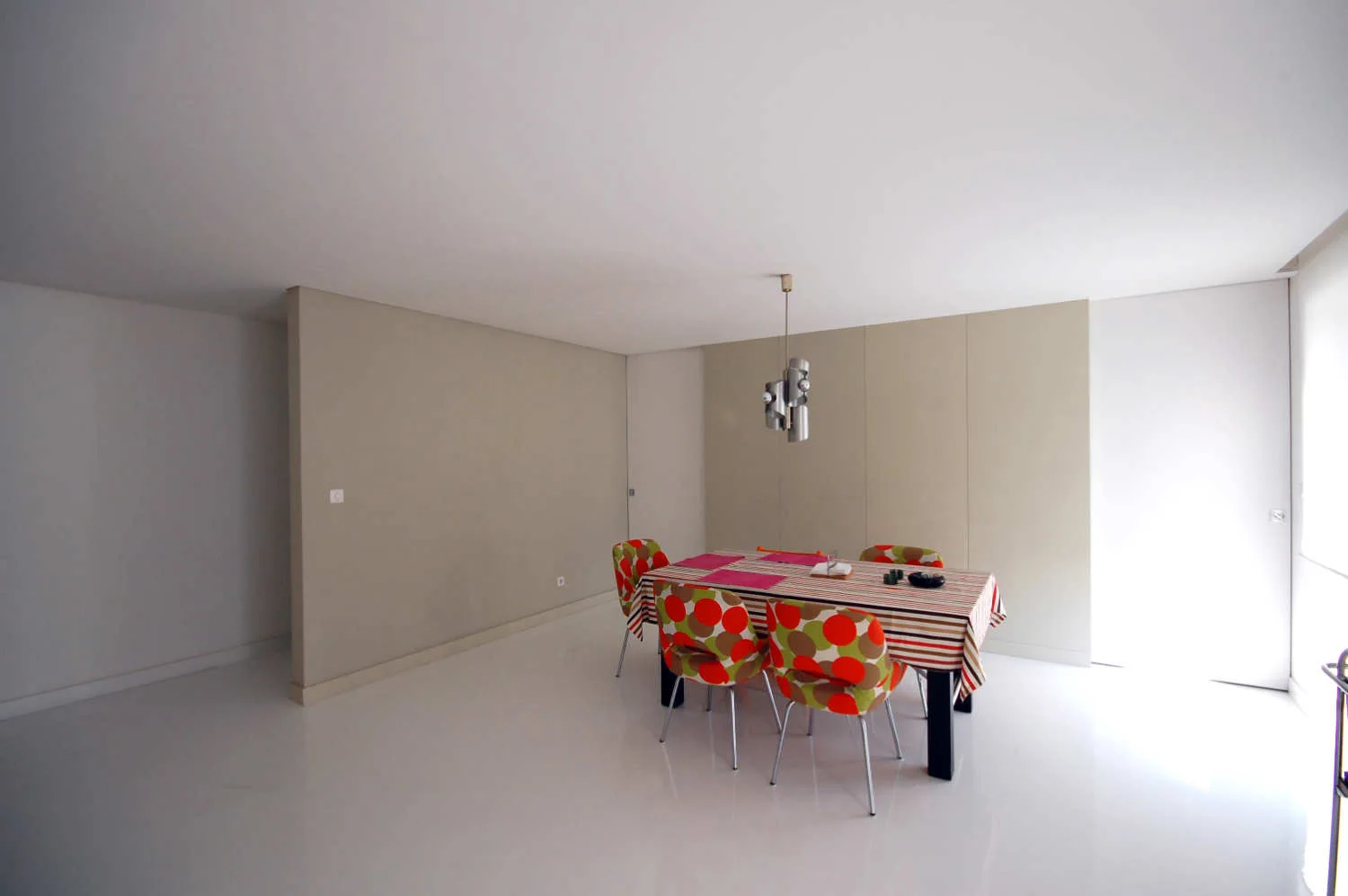 A minimalist dining table with colorful patterned chairs is set under a silver pendant light in a bright room with beige panels.