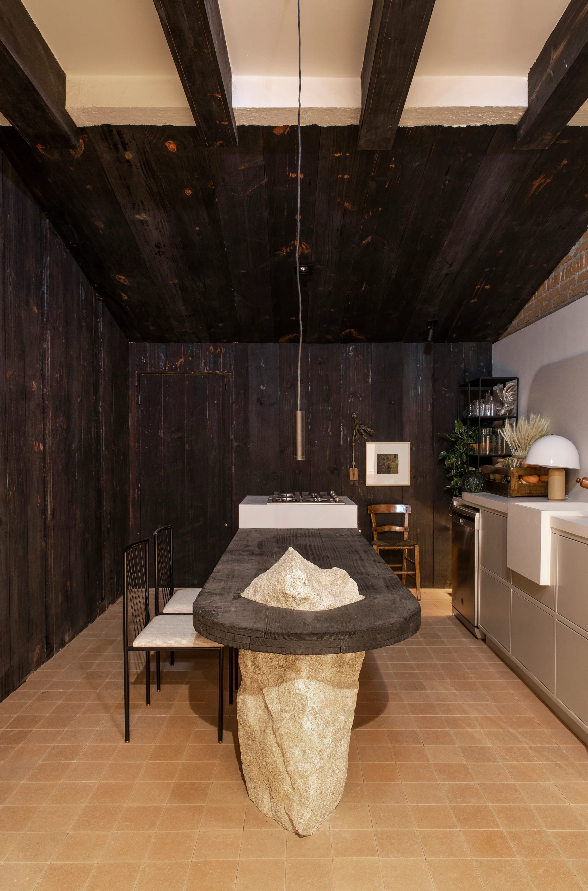 A kitchen and dining space features a central wooden table supported by a large raw stone block, surrounded by dark wood walls and beige tile floors.