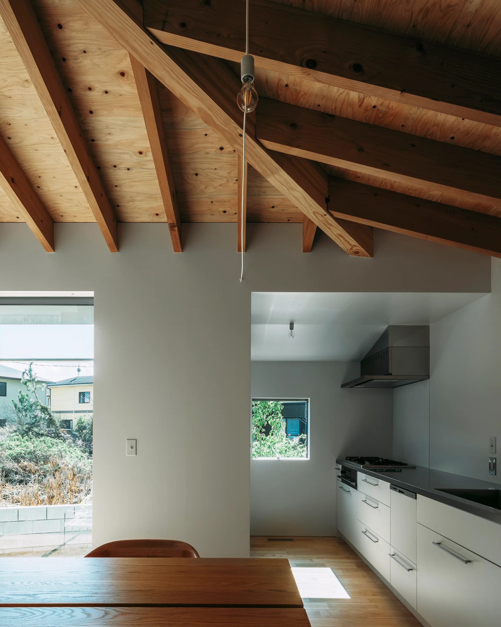 A minimalist kitchen features a high vaulted ceiling with exposed plywood beams, white cabinets, and large windows looking outside.