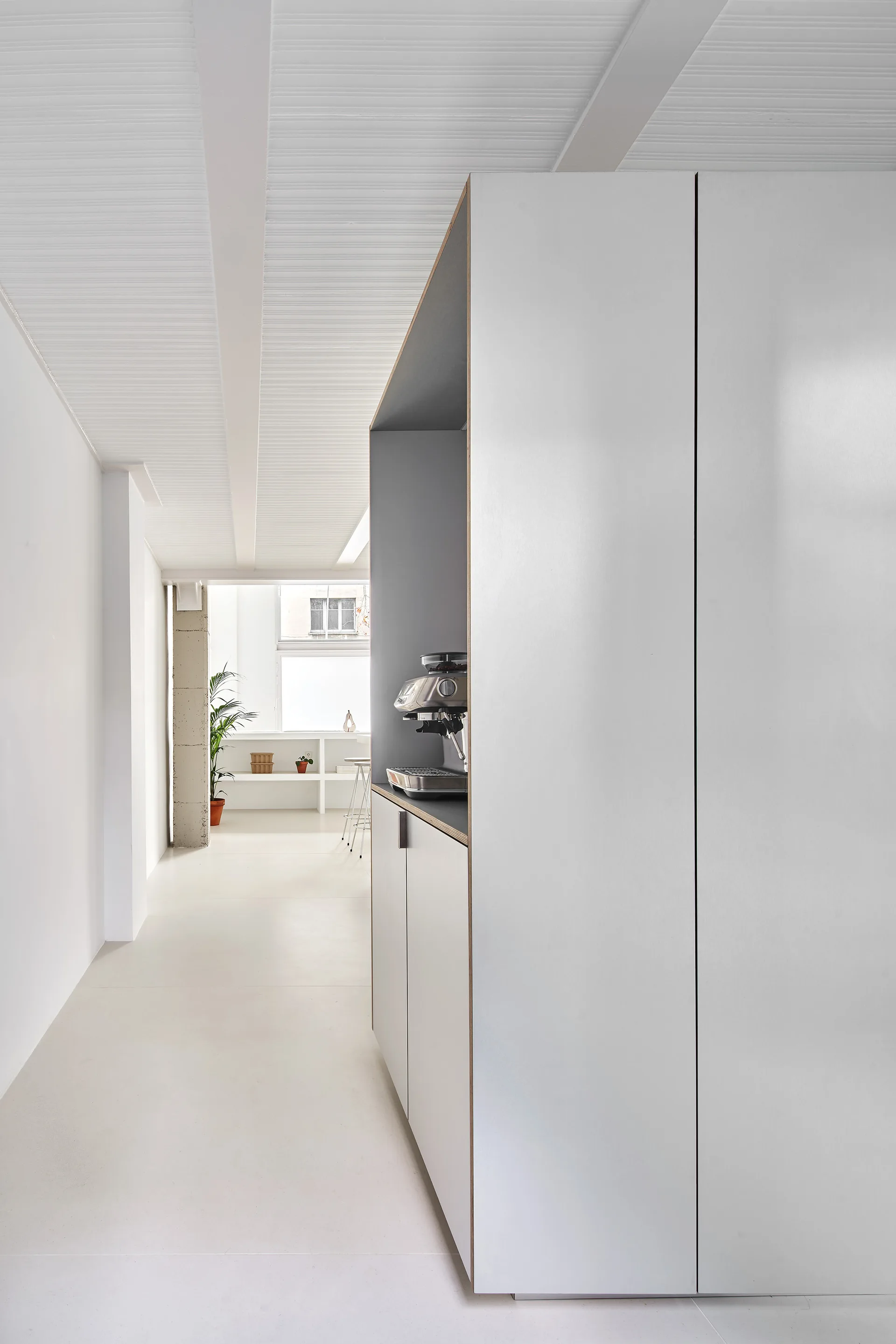 A large, minimalist white cabinet unit with a recessed coffee niche sits in a bright room featuring ceiling beams and a white floor.