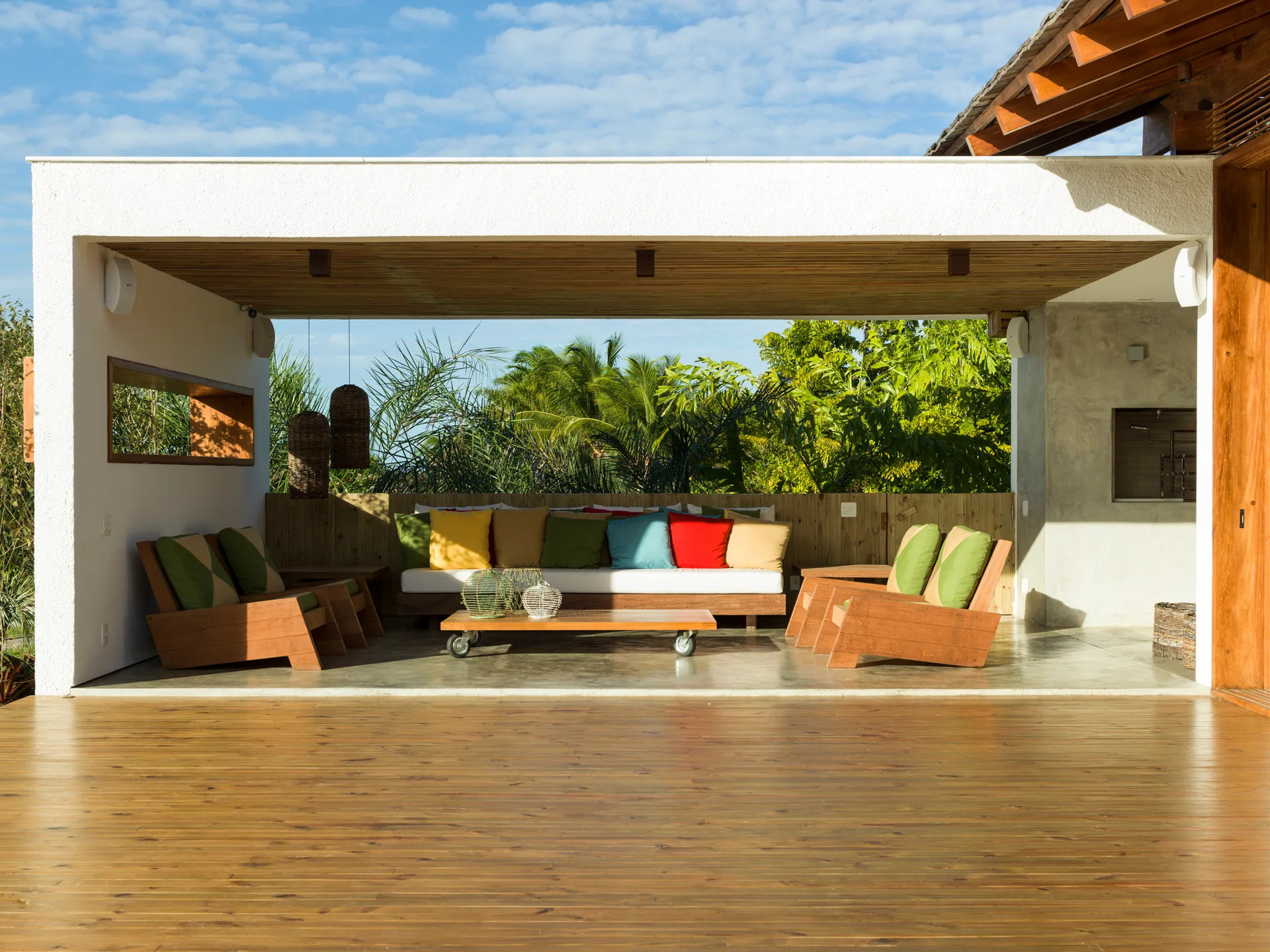 A covered wooden patio with a slatted ceiling features an outdoor sofa with vibrant pillows and armchairs overlooking a tropical garden.