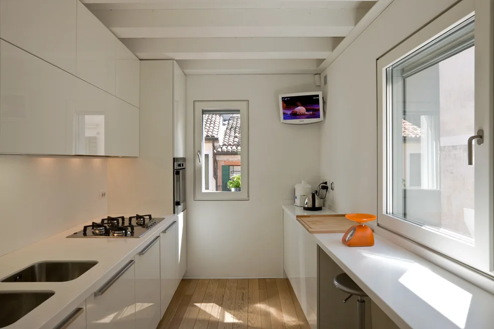 A narrow galley kitchen features glossy white cabinets, light wood flooring, exposed ceiling beams, and a stainless steel sink.