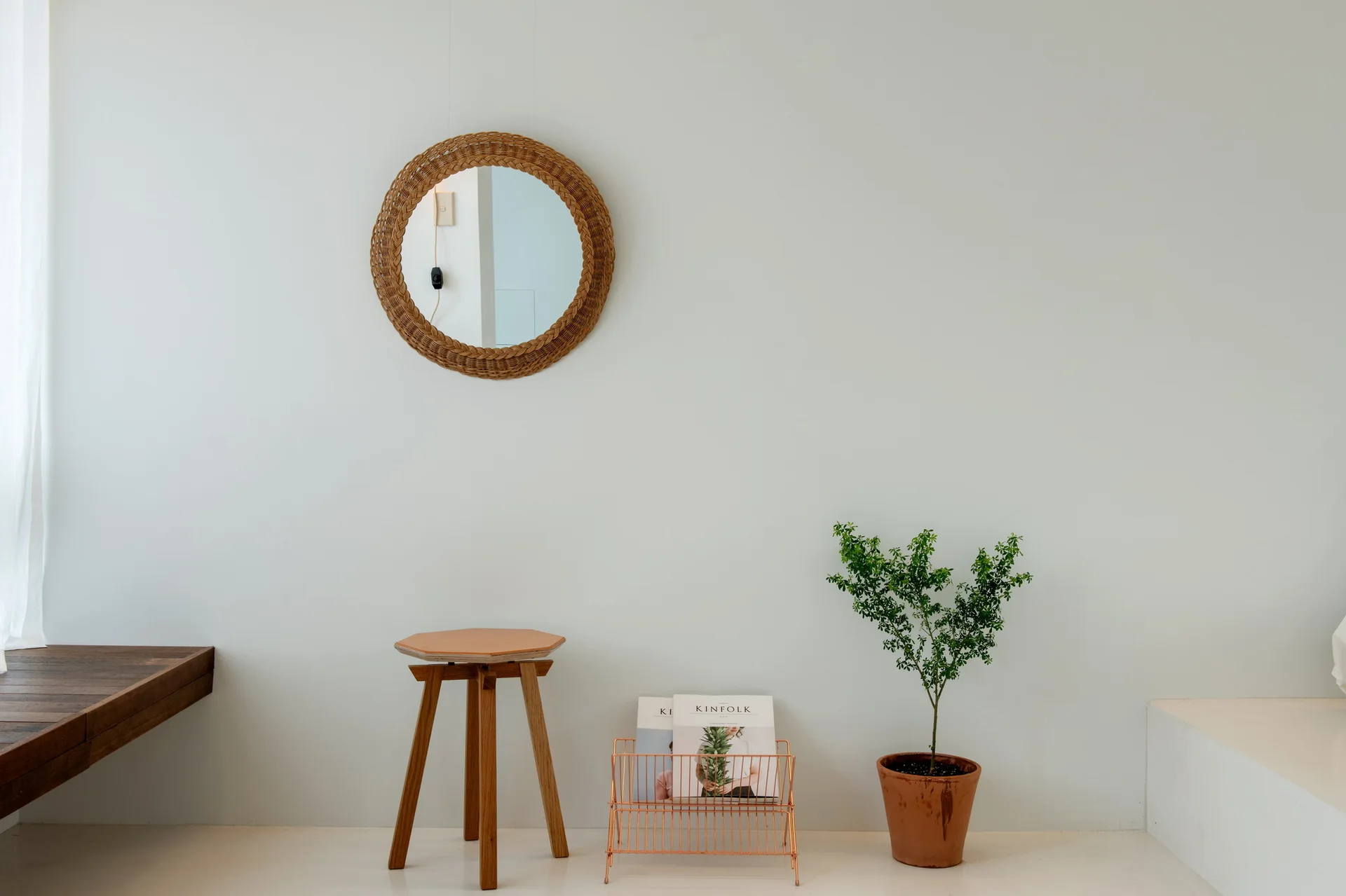 A circular rattan mirror hangs on a light gray wall above a wooden stool, copper magazine rack, and a small potted plant.