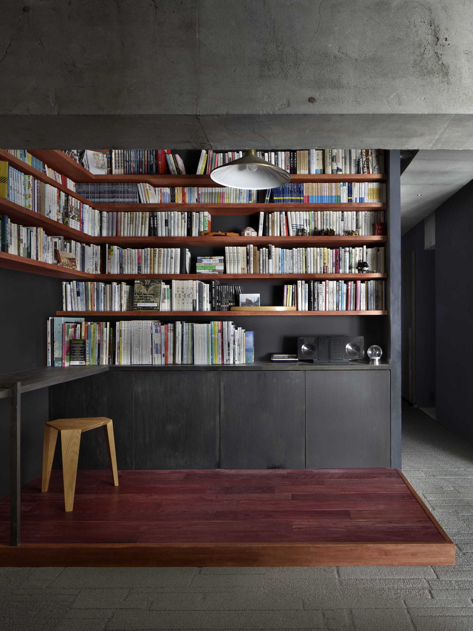 A minimalist home library with floor-to-ceiling wooden bookshelves, dark charcoal walls, a raised wooden platform floor, and an exposed concrete ceiling.