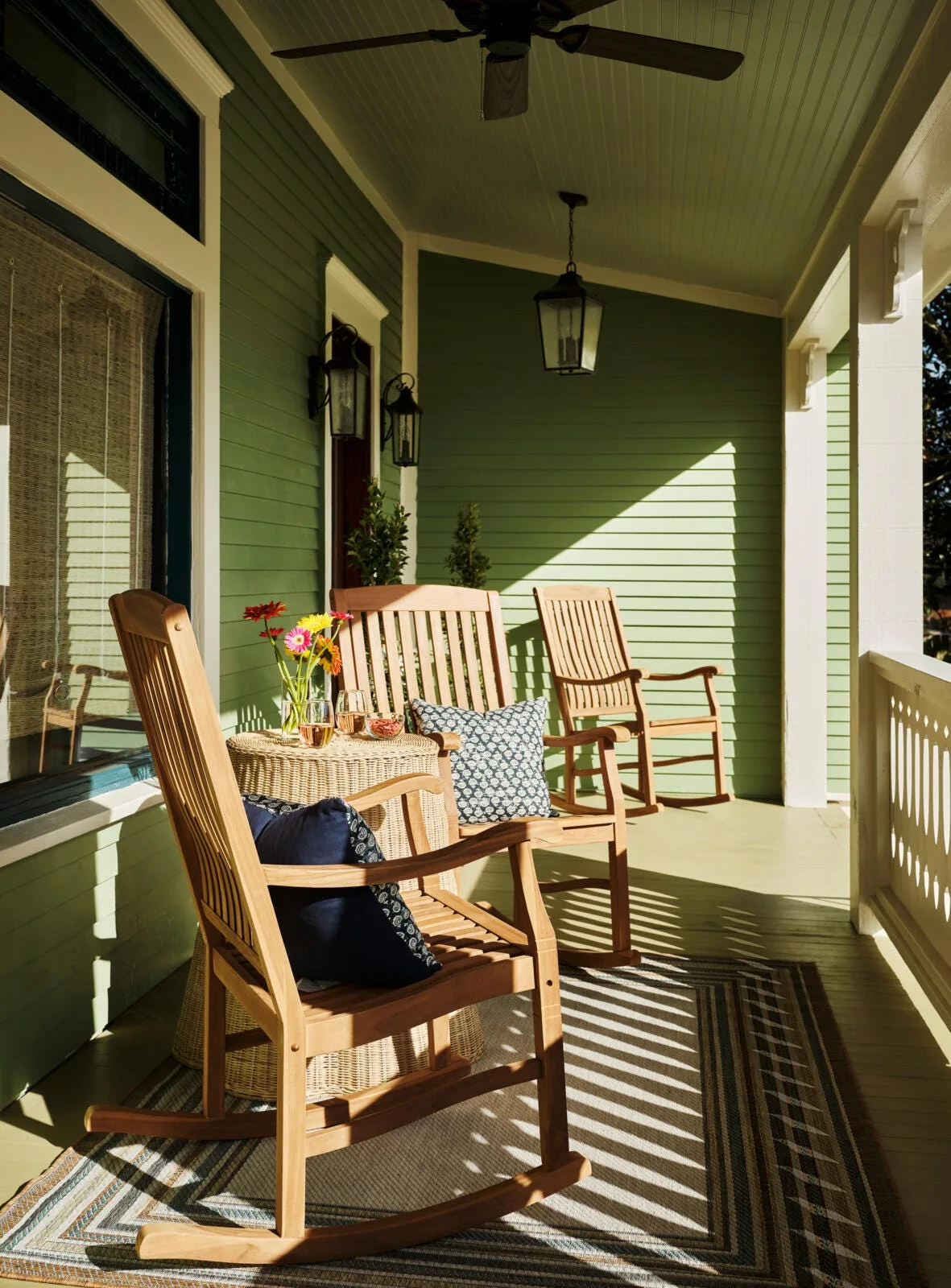Three wooden rocking chairs sit on a covered green porch with matching siding, a wicker side table, and a striped area rug.