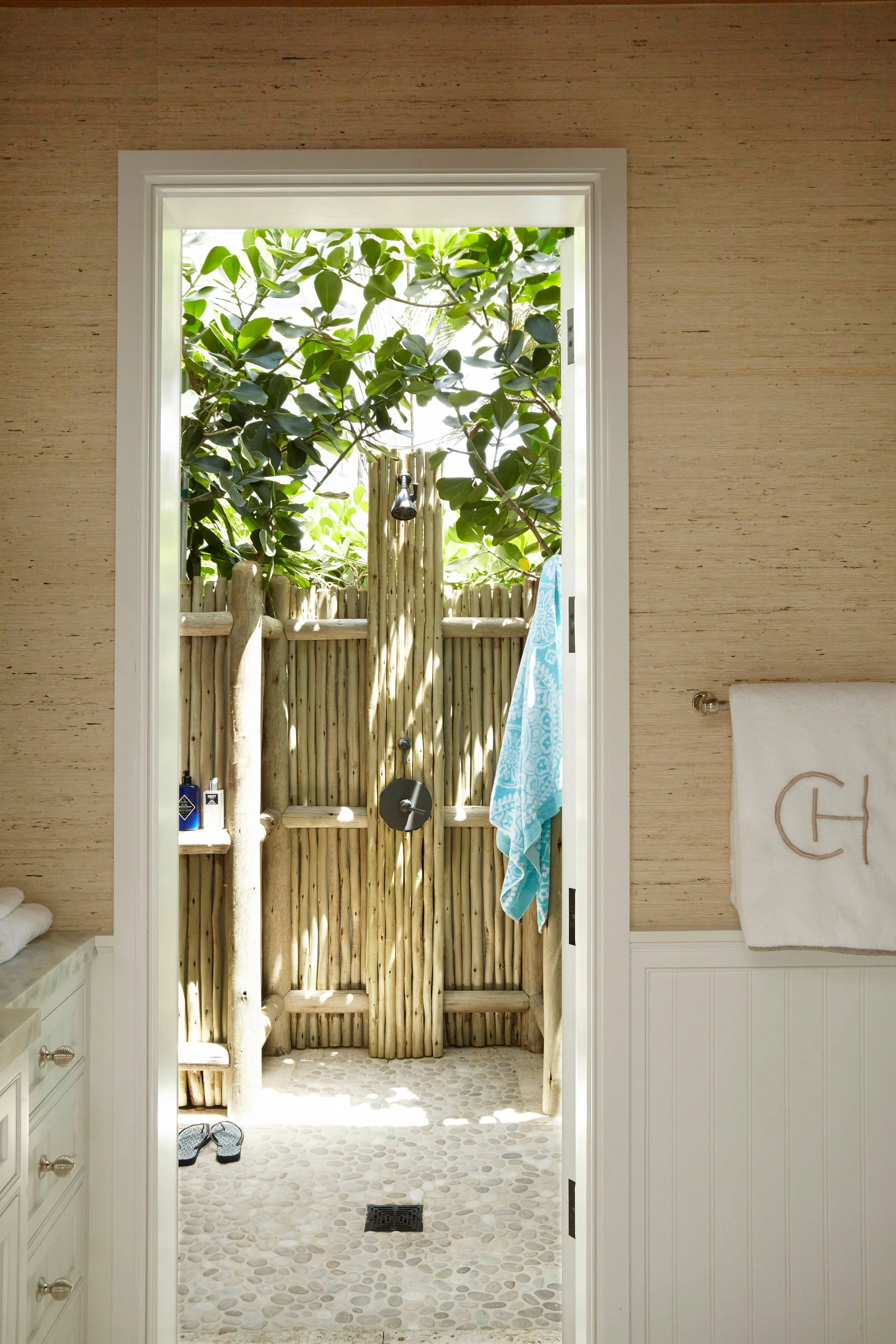 A view through a white door frame from a bathroom with textured wallpaper to an outdoor shower built with vertical wood poles and a pebble floor.