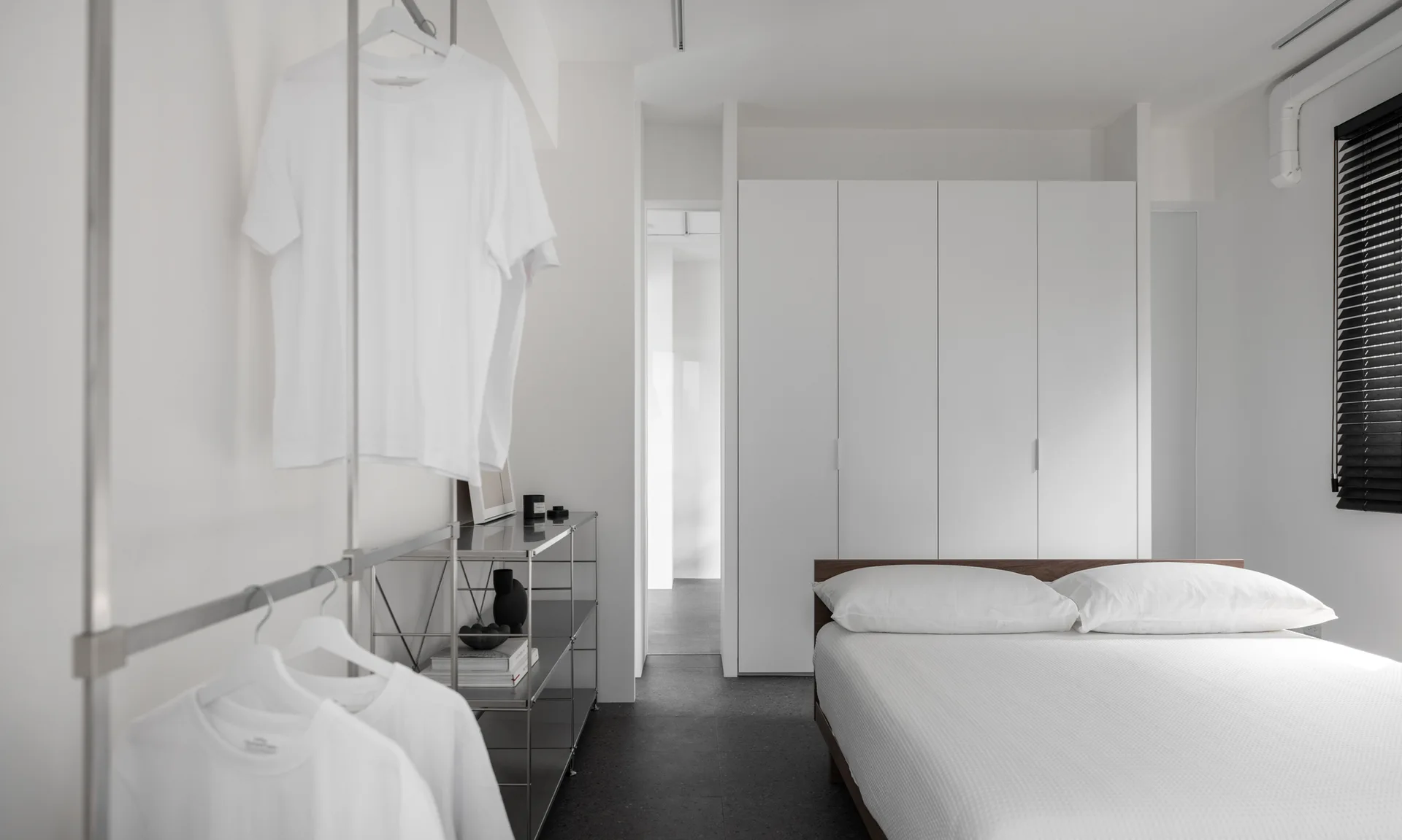 A minimalist white bedroom featuring a wood bed frame, floor-to-ceiling built-in wardrobes, and a metal modular clothing rack with hanging white shirts.