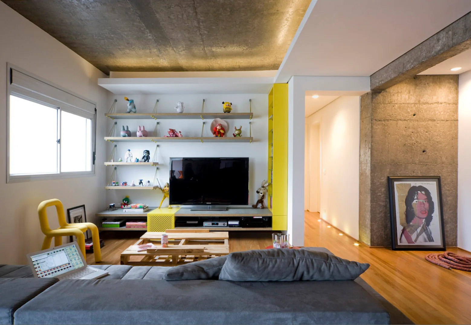 An industrial living room with a grey sofa, an exposed concrete ceiling, open shelving displaying figurines, and bright yellow accent furniture.