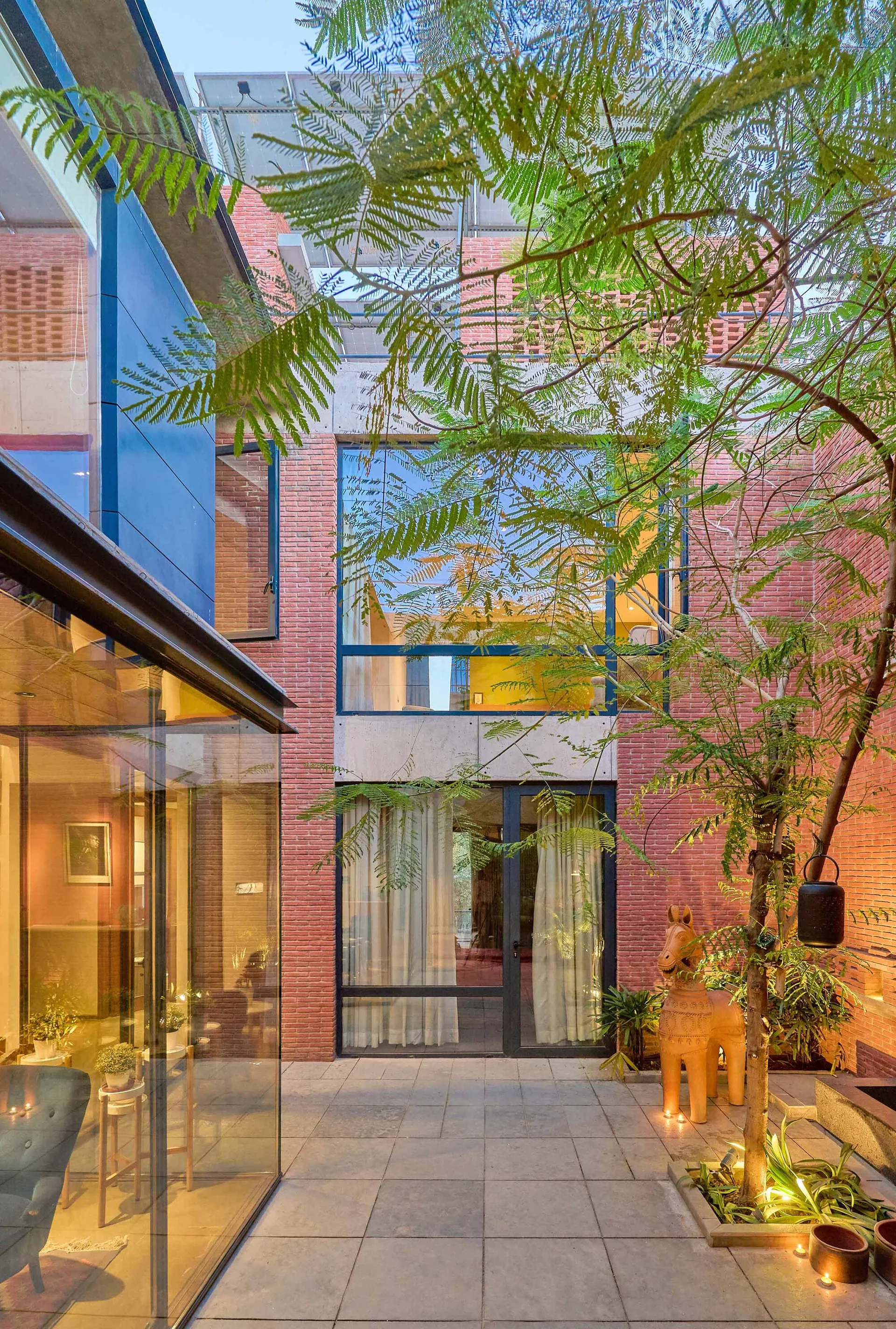 A modern paved courtyard features red brick walls, large glass windows showing interior spaces, a central tree, and a horse sculpture.