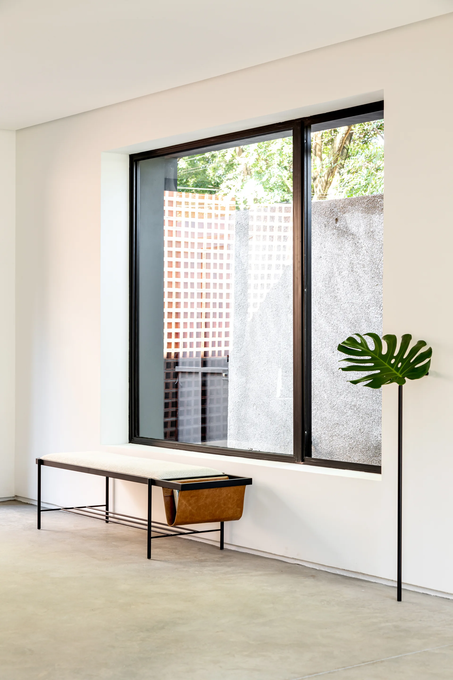 A minimalist black metal bench with a light cushion and leather magazine sling sits below a large window on concrete flooring.