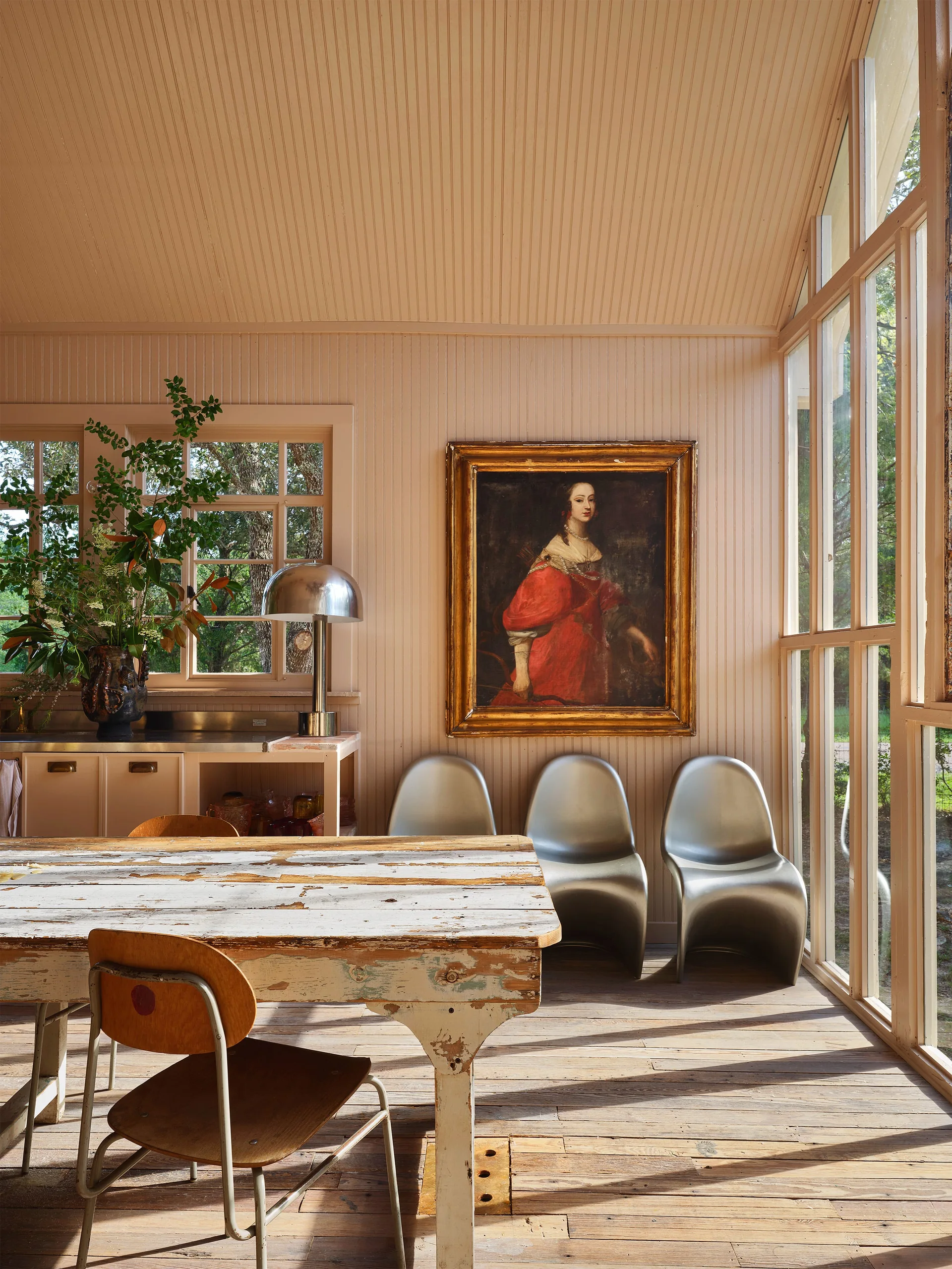 A rustic wooden dining table sits in a light pink paneled dining room beside three silver metallic chairs and a large framed portrait.