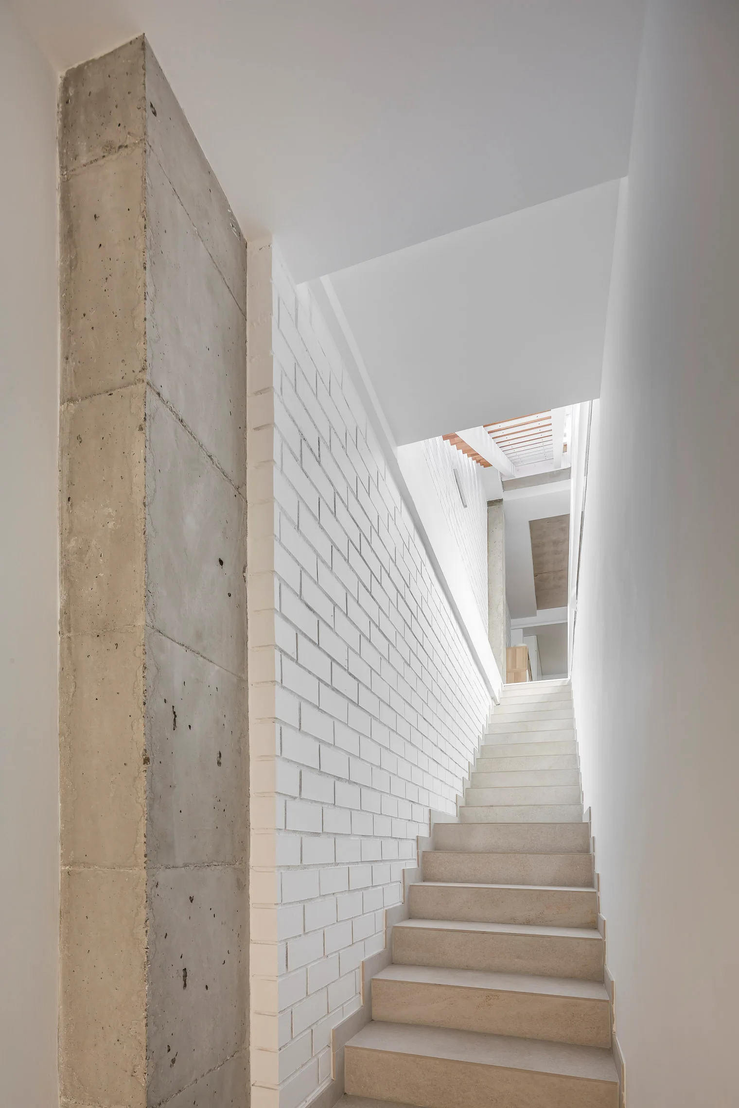 A straight indoor staircase with light stone steps rises between a white painted brick wall and a raw concrete column under natural light.