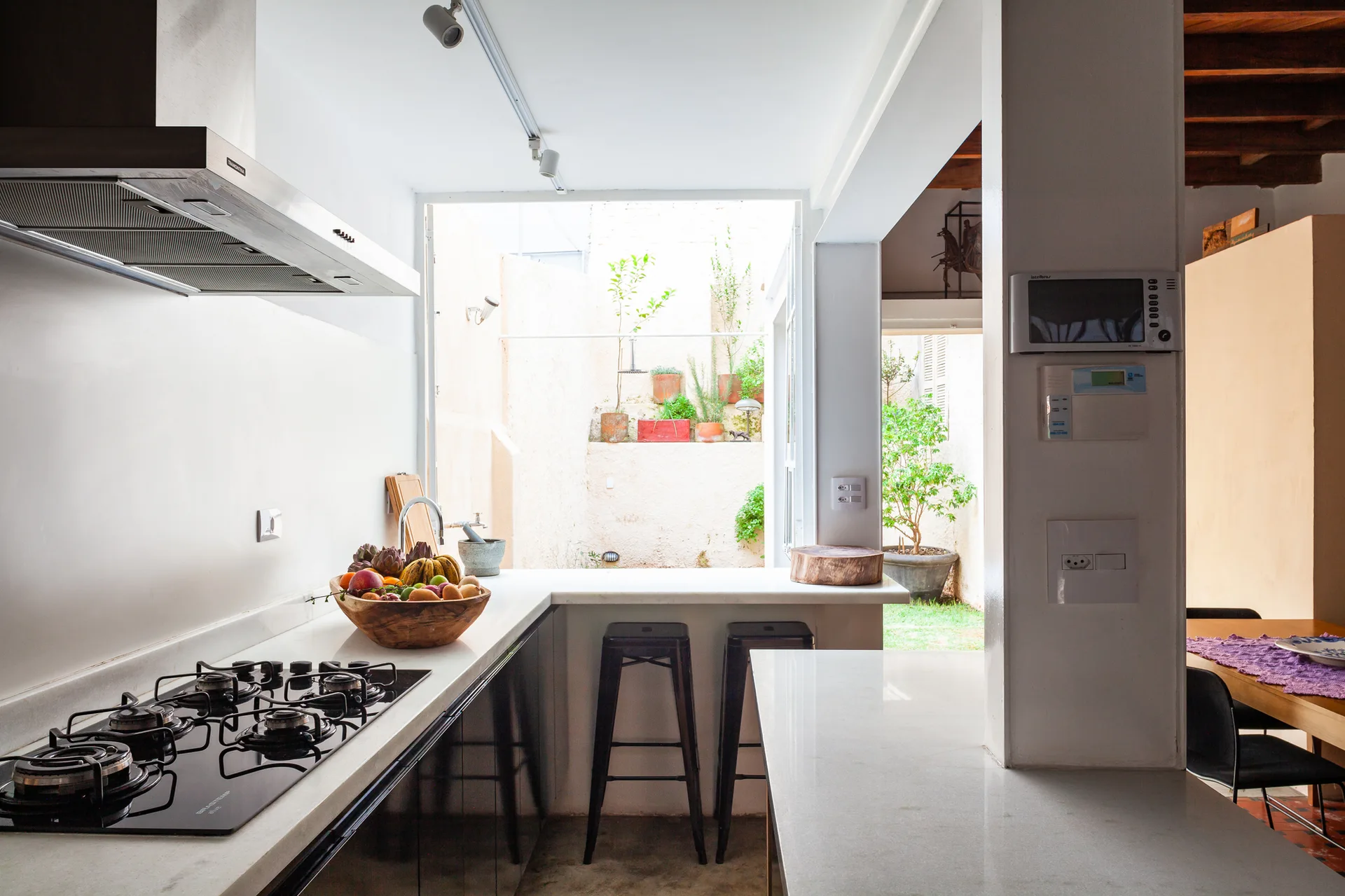 A minimalist kitchen features a white countertop with a black stovetop, metal bar stools, and a wide opening looking onto an outdoor courtyard.