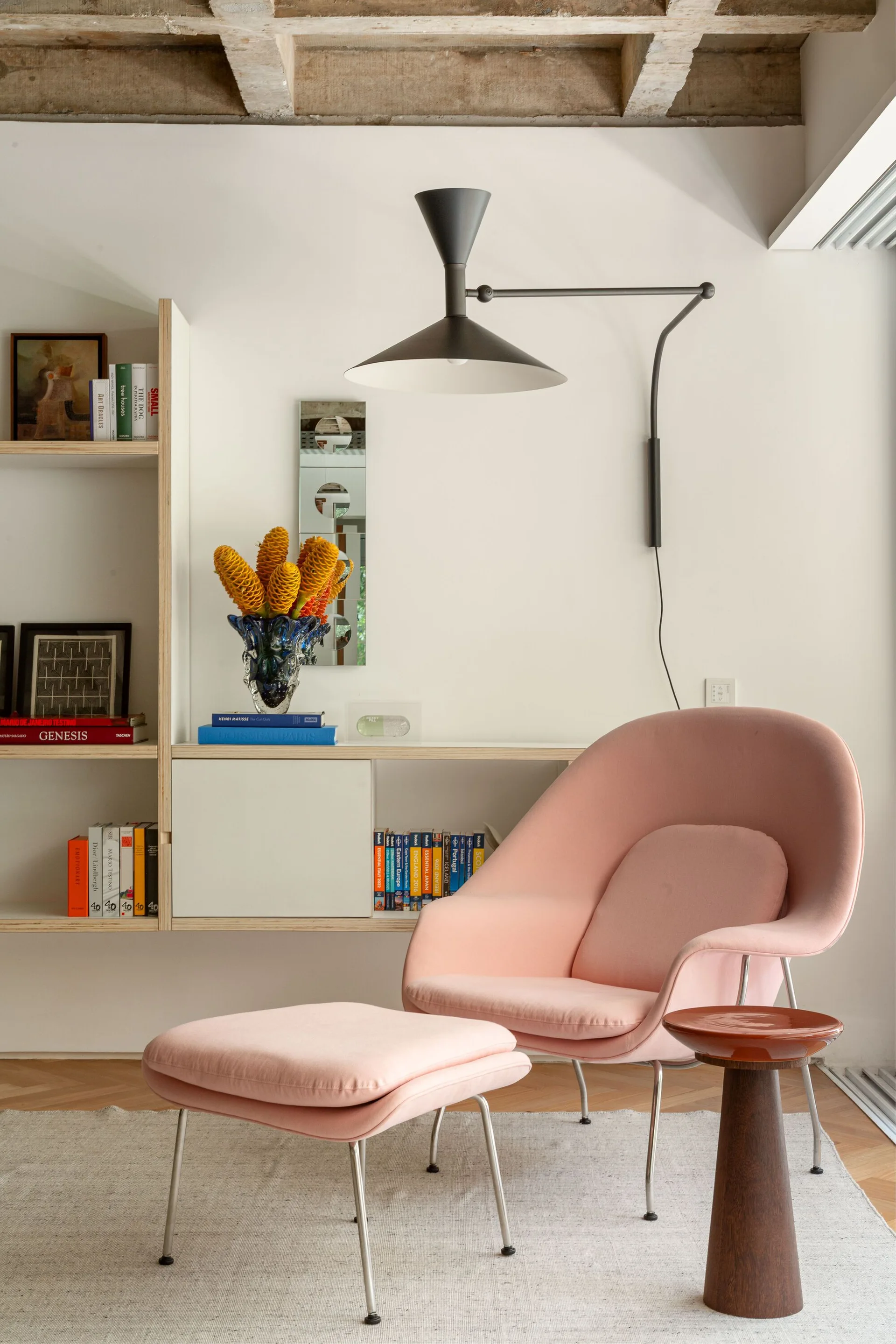 A reading nook features a light pink lounge chair and ottoman set on a light rug next to a light wood shelving unit filled with books.