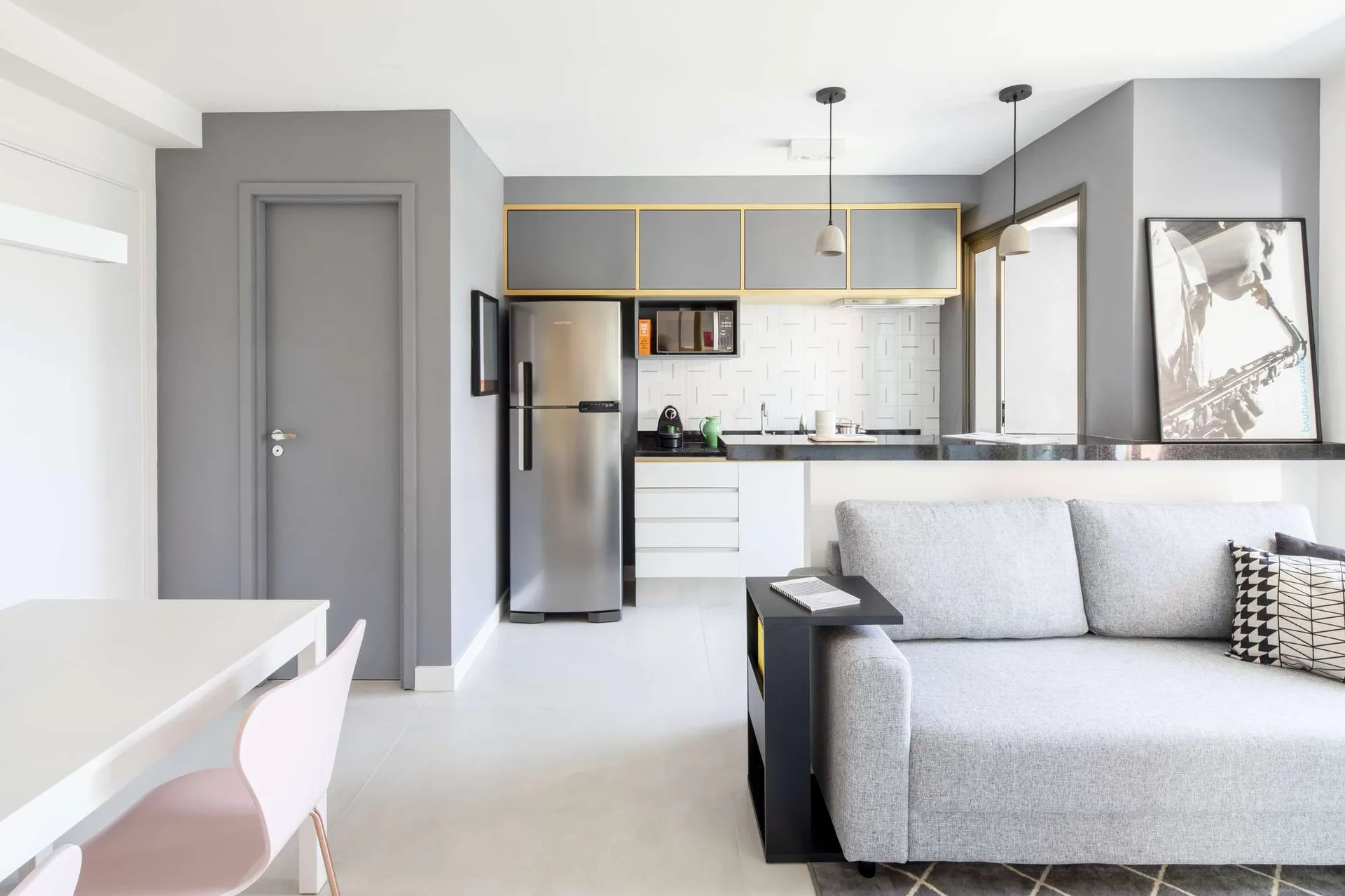A modern grey and white open concept apartment showing a grey sofa in the living area and minimalist kitchen cabinets with gold trim.