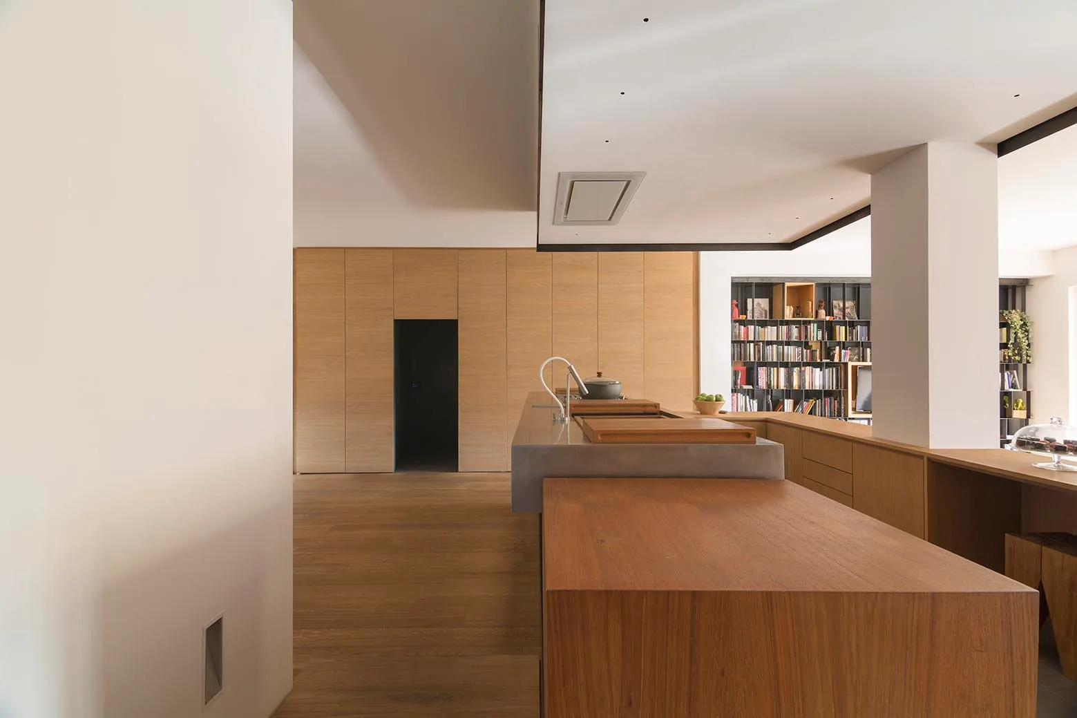 A minimalist kitchen features flat-panel oak cabinets, a dual-material wood and concrete island, and a recessed library in the background.