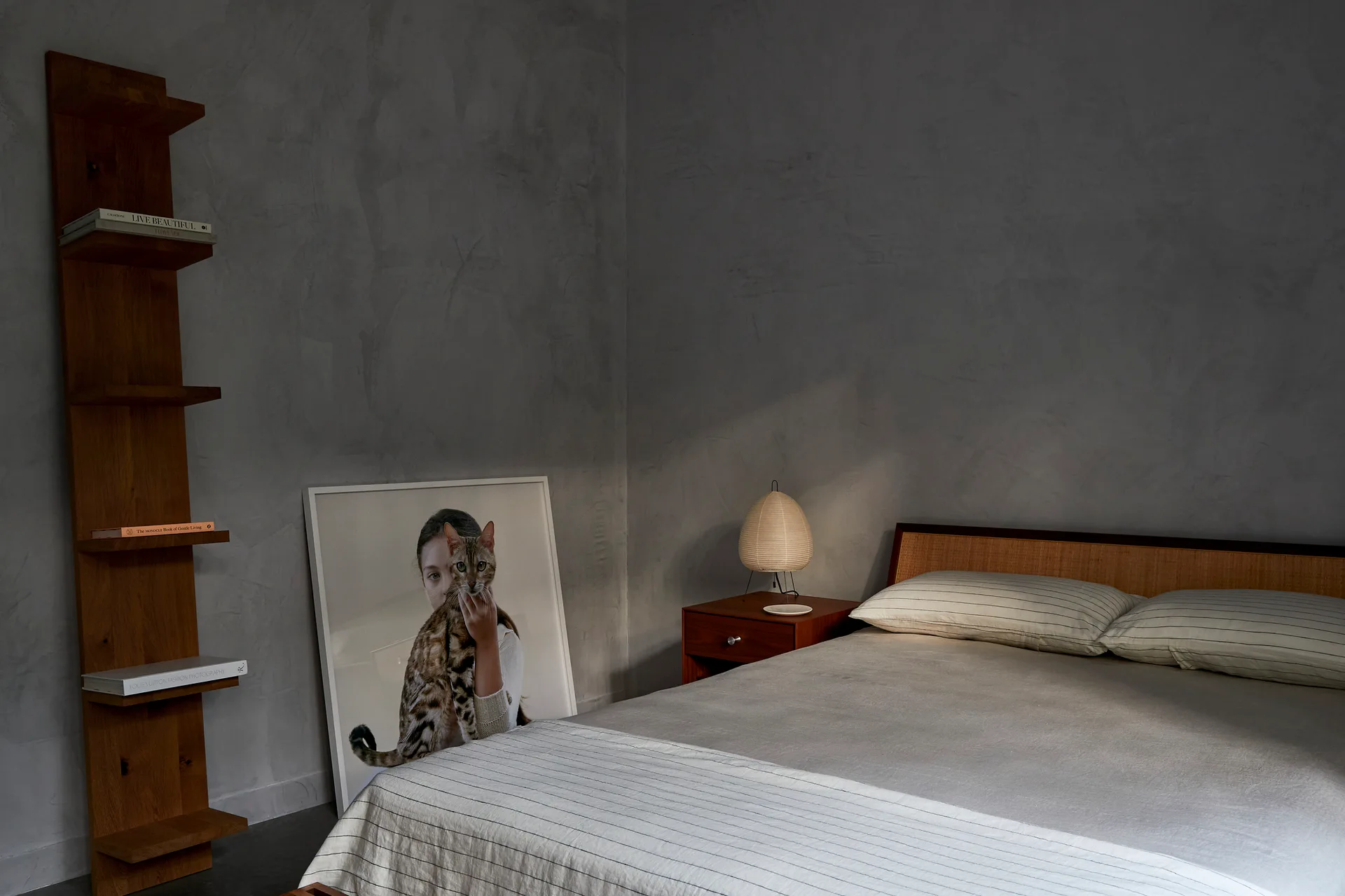 A minimalist bedroom features a low wooden bed, a bedside table lamp, a leaning framed photo, and a tall wood shelf against textured grey plaster walls.