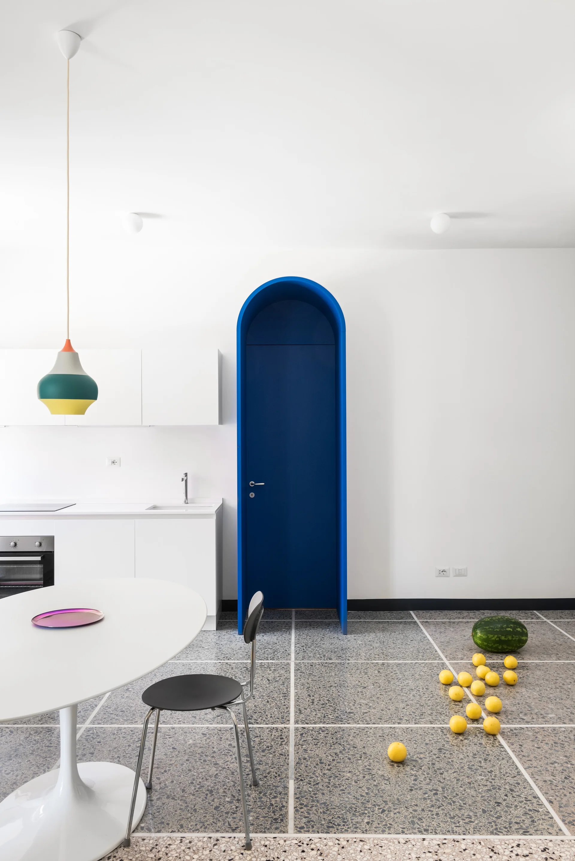 A minimalist white kitchen features a bright blue arched entryway, white cabinets, a round dining table, and grey speckled terrazzo tile floors.