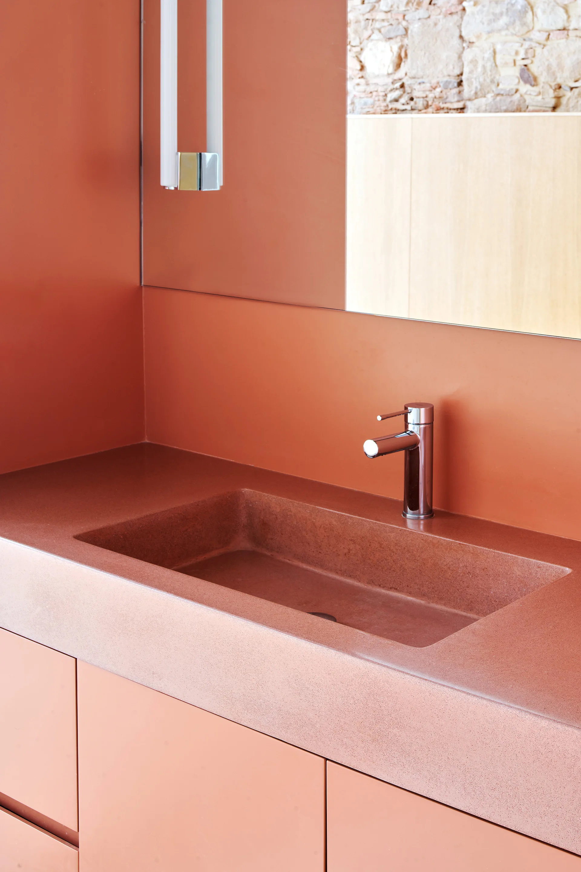 A minimalist terracotta bathroom vanity features a speckled integrated sink, a polished chrome faucet, and a large mirror reflecting a stone wall.