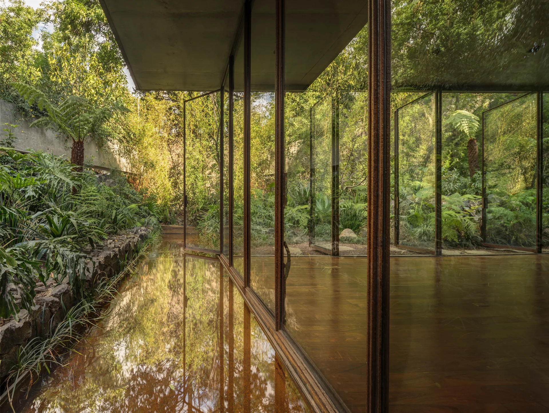 An interior room with polished wood flooring features a floor-to-ceiling glass wall overlooking a lush green garden and a reflective outdoor pool.
