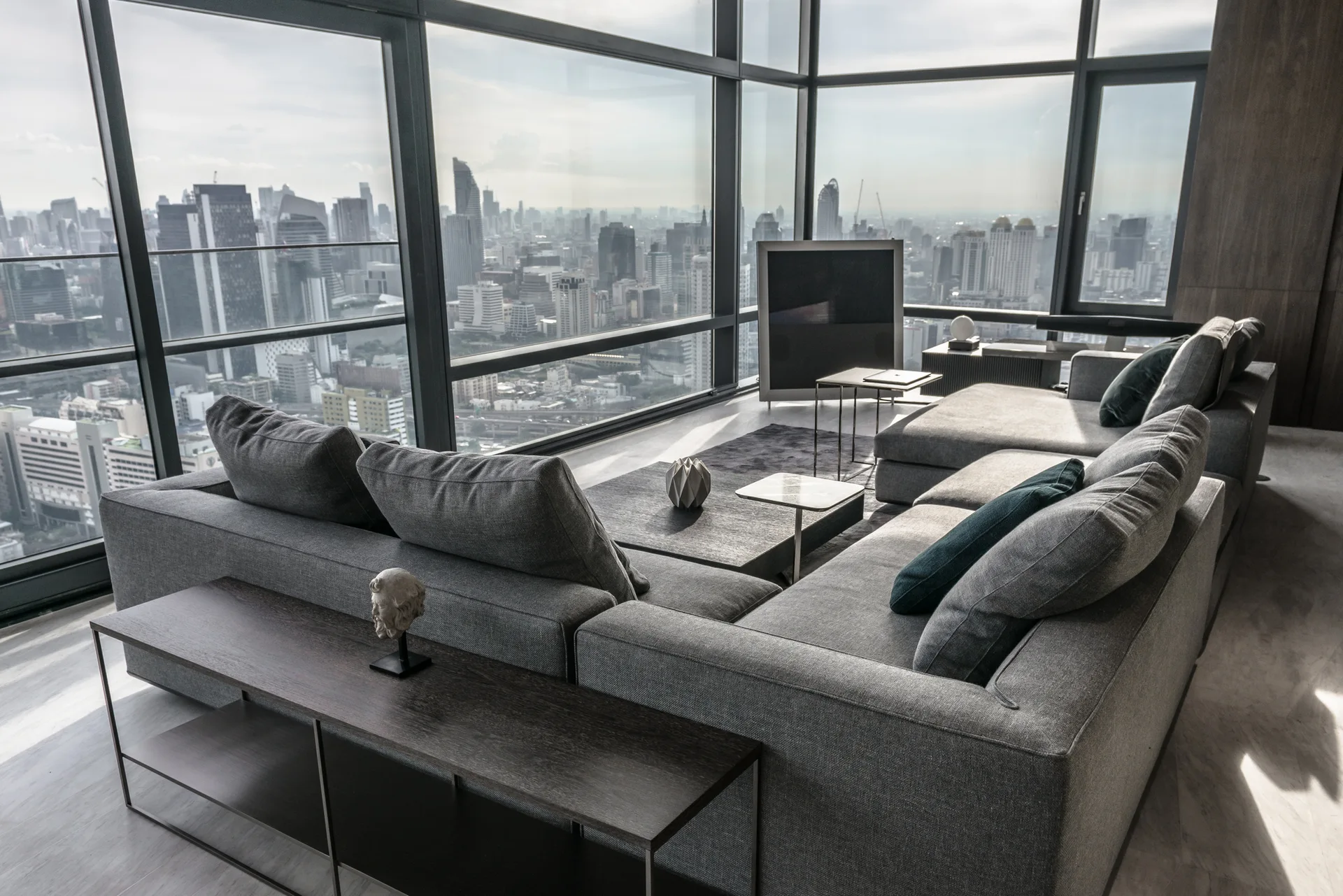 Two grey sectional sofas frame a low coffee table in a high-rise living room surrounded by panoramic corner windows with city views.