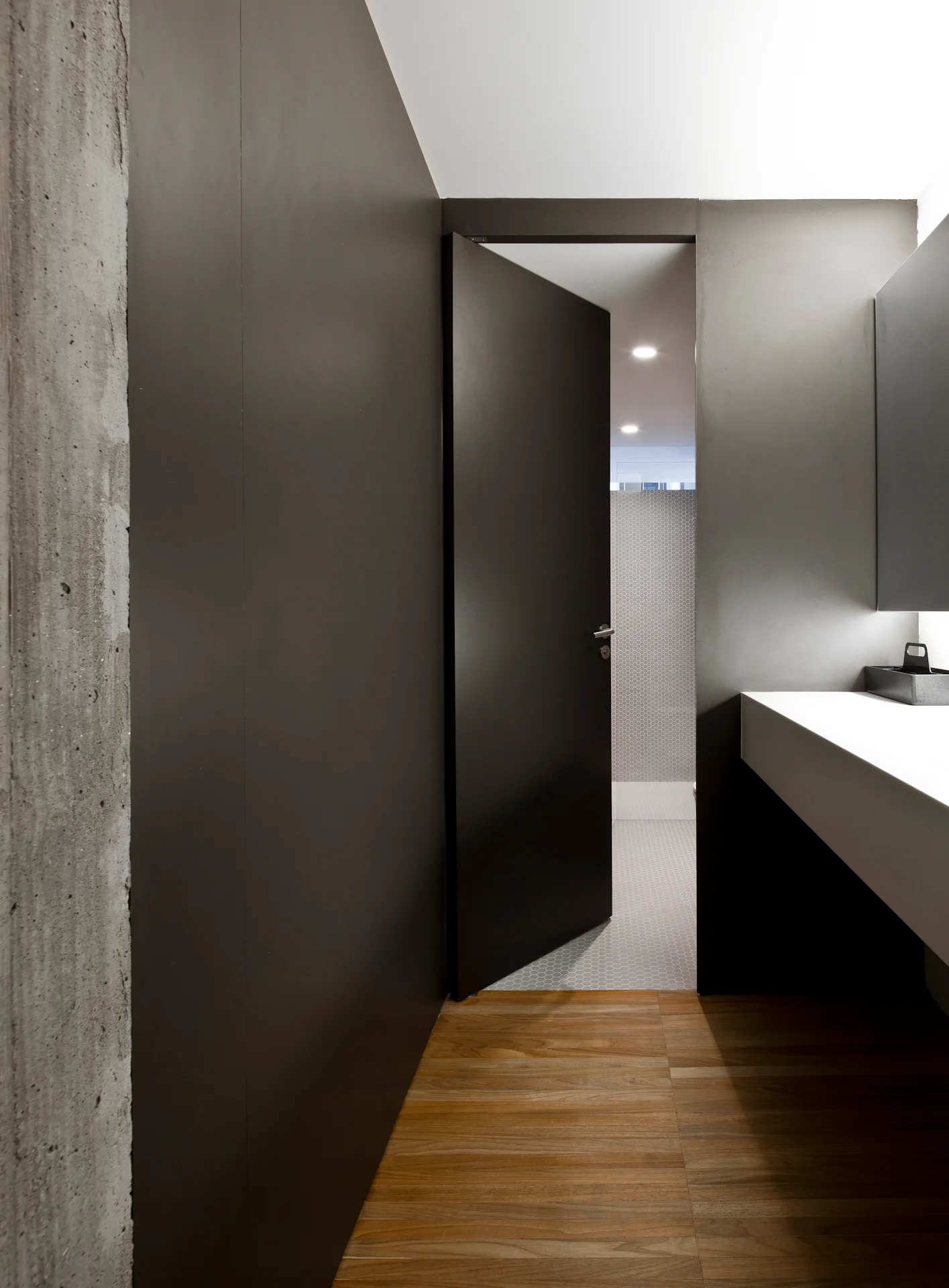 A modern bathroom entryway features wood plank flooring, charcoal walls with a matching door, an exposed concrete pillar, and a white floating vanity.