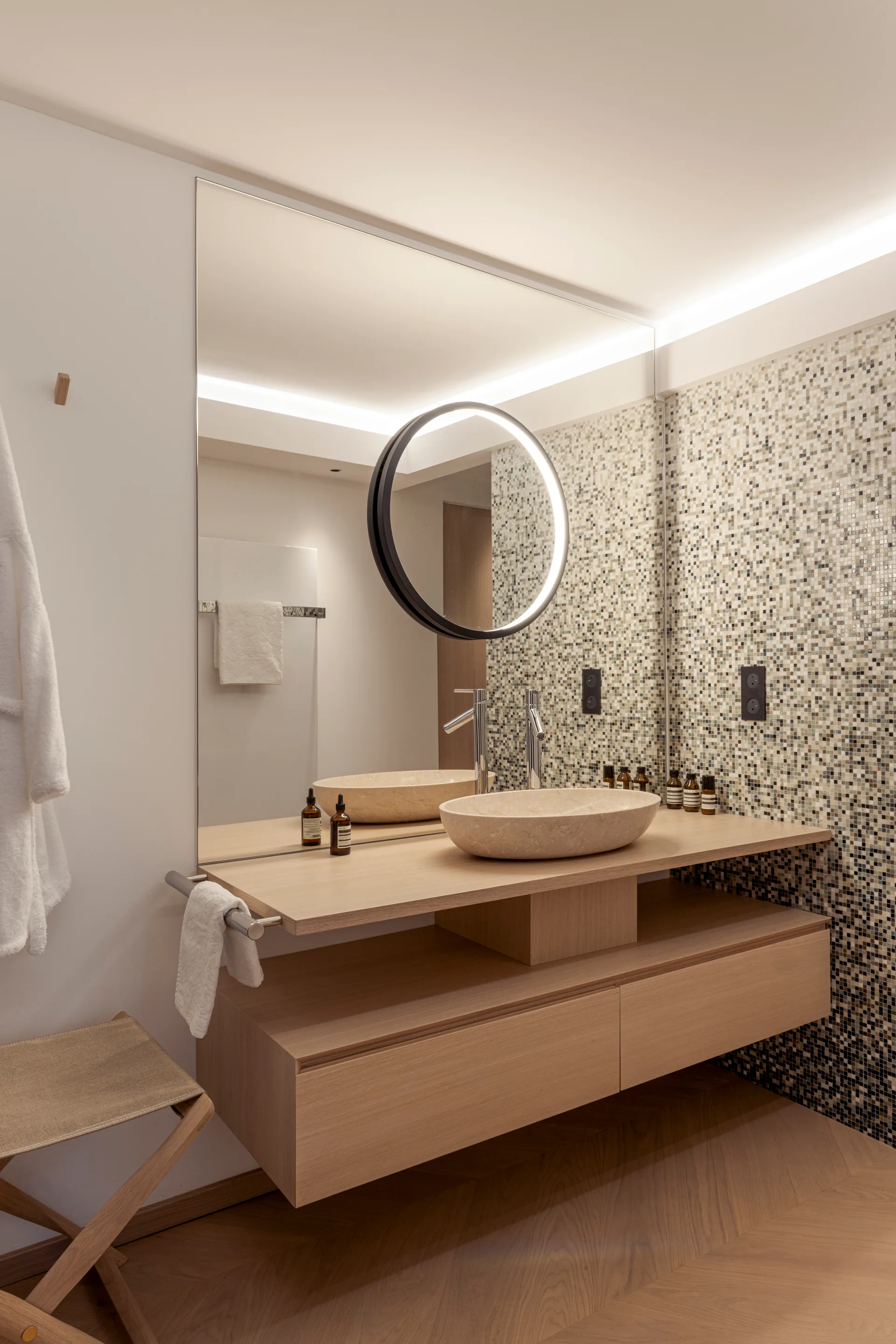 A modern bathroom features a floating oak vanity with an oval stone vessel sink, a mosaic tiled wall, and a large mirror with a circular illuminated inset.
