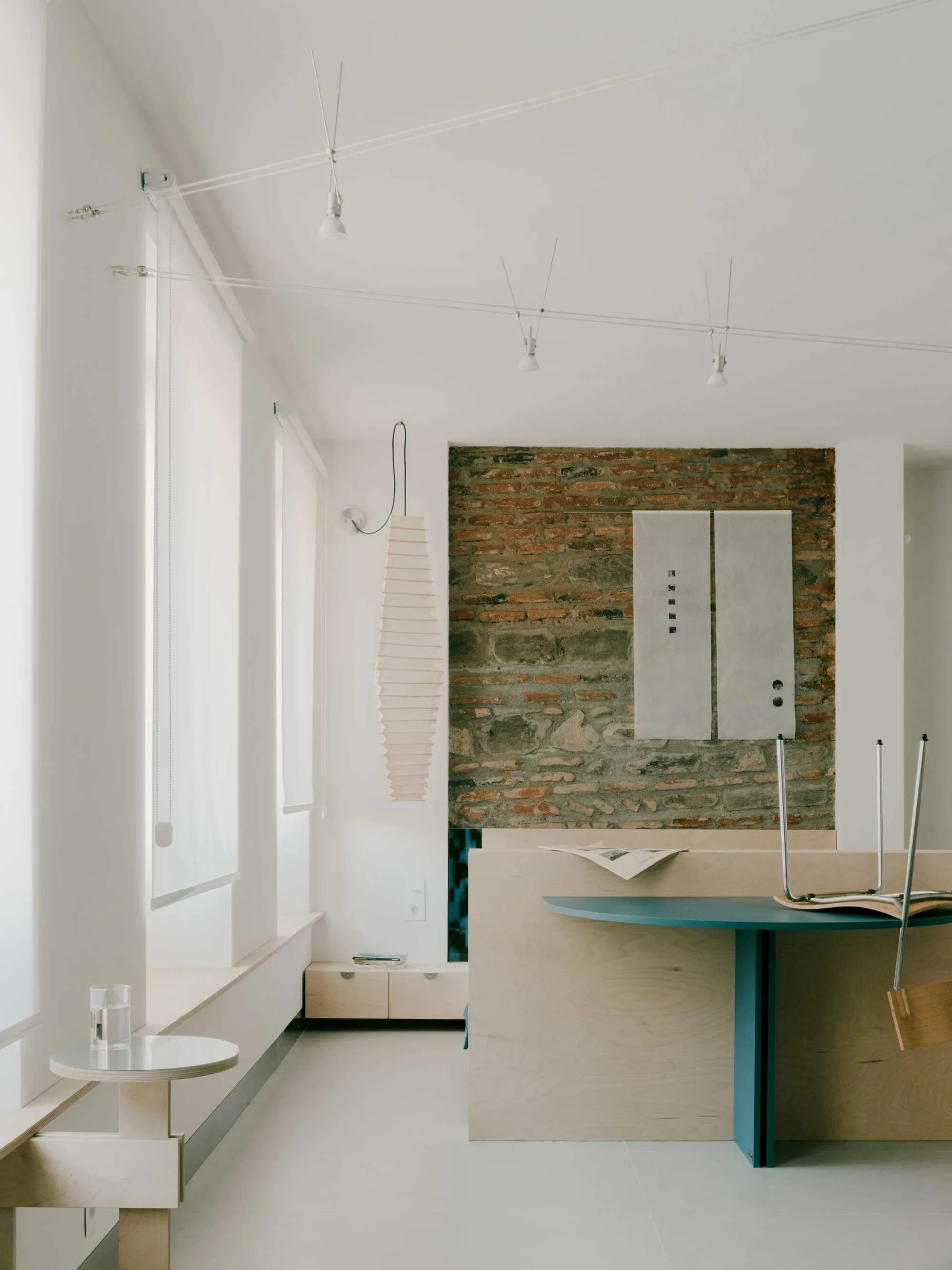 A minimalist room featuring a blue round table, light wood partitions, an exposed brick wall, and overhead cable lighting.