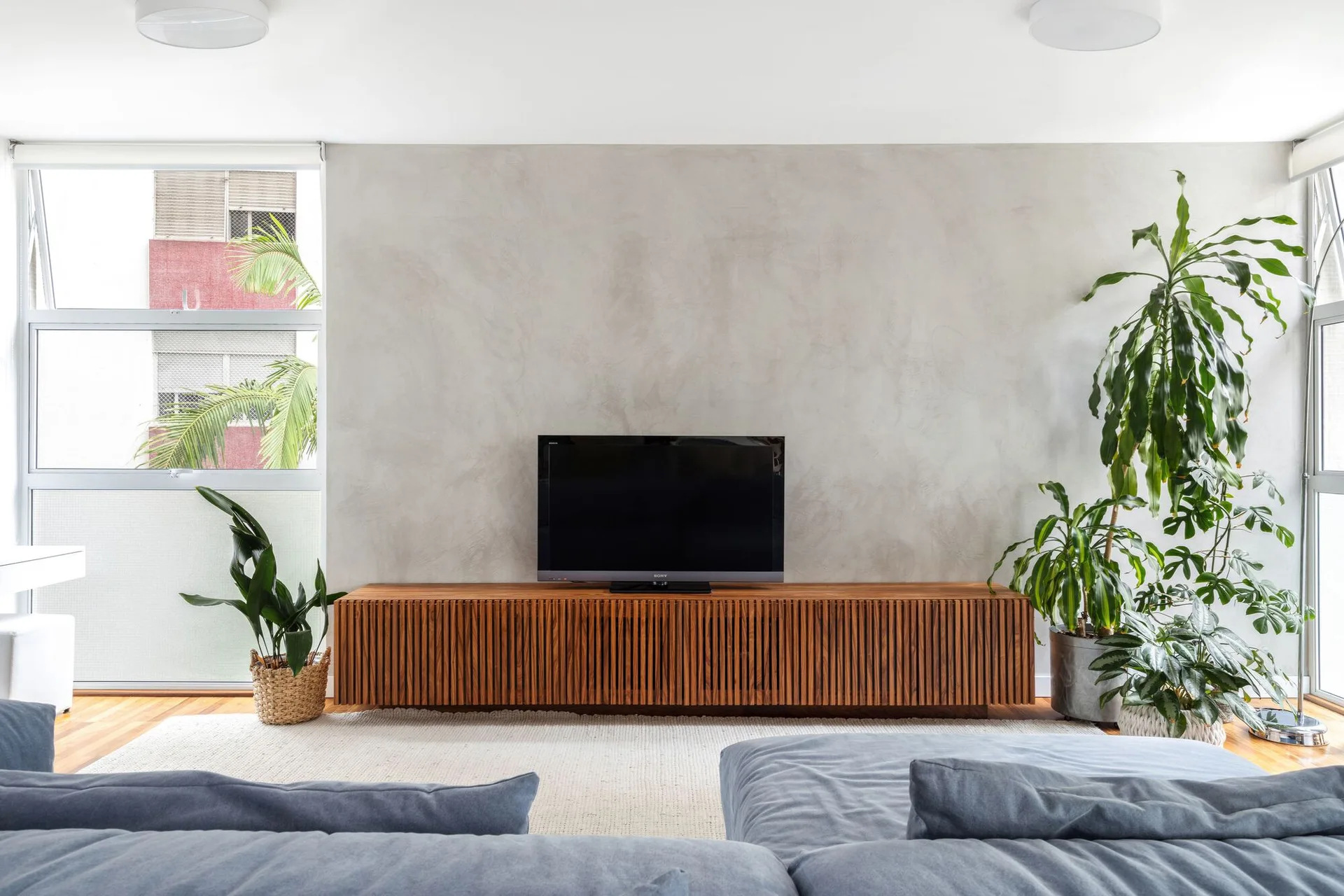 A slatted wood TV console stands against a textured gray plaster wall flanked by tall windows and potted green plants in a living room.