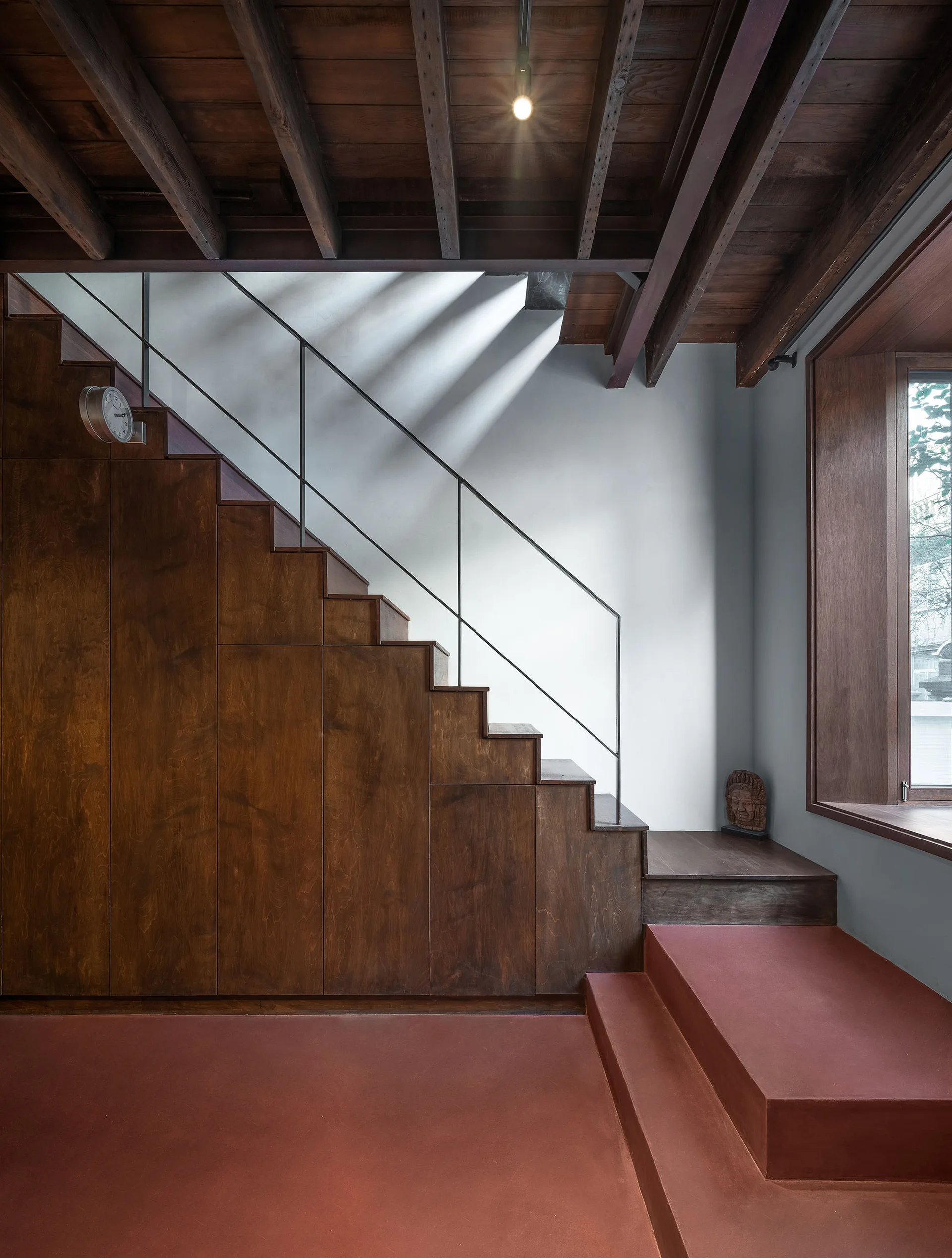 A modern wooden staircase featuring dark wood cabinetry built underneath, exposed timber ceiling beams, and red terracotta concrete steps.