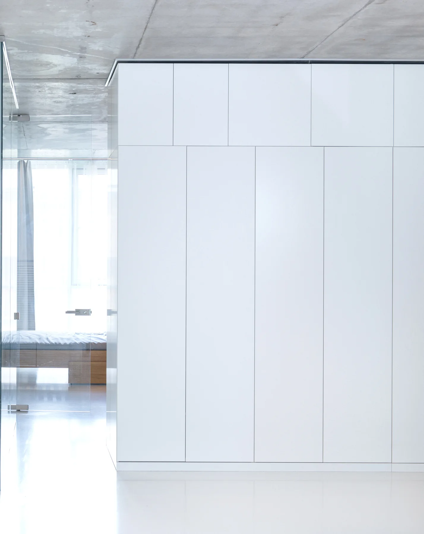 A minimalist bedroom entry features a tall, seamless white built-in wardrobe beneath a textured grey concrete ceiling with a clear glass partition.