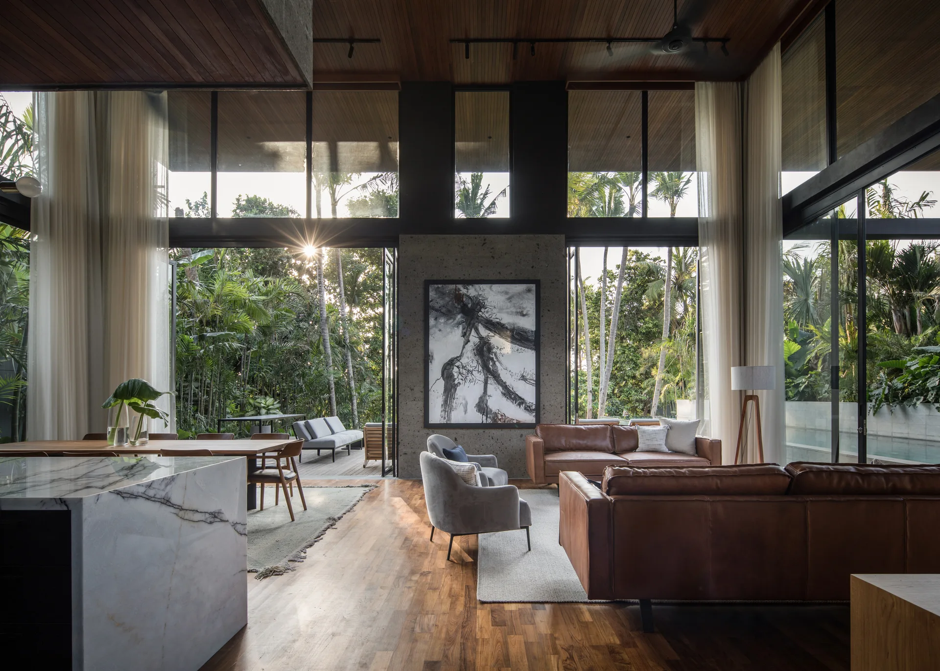 An open living room featuring wood plank ceilings, leather sofas, grey armchairs, and floor-to-ceiling glass walls looking out to a tropical garden.