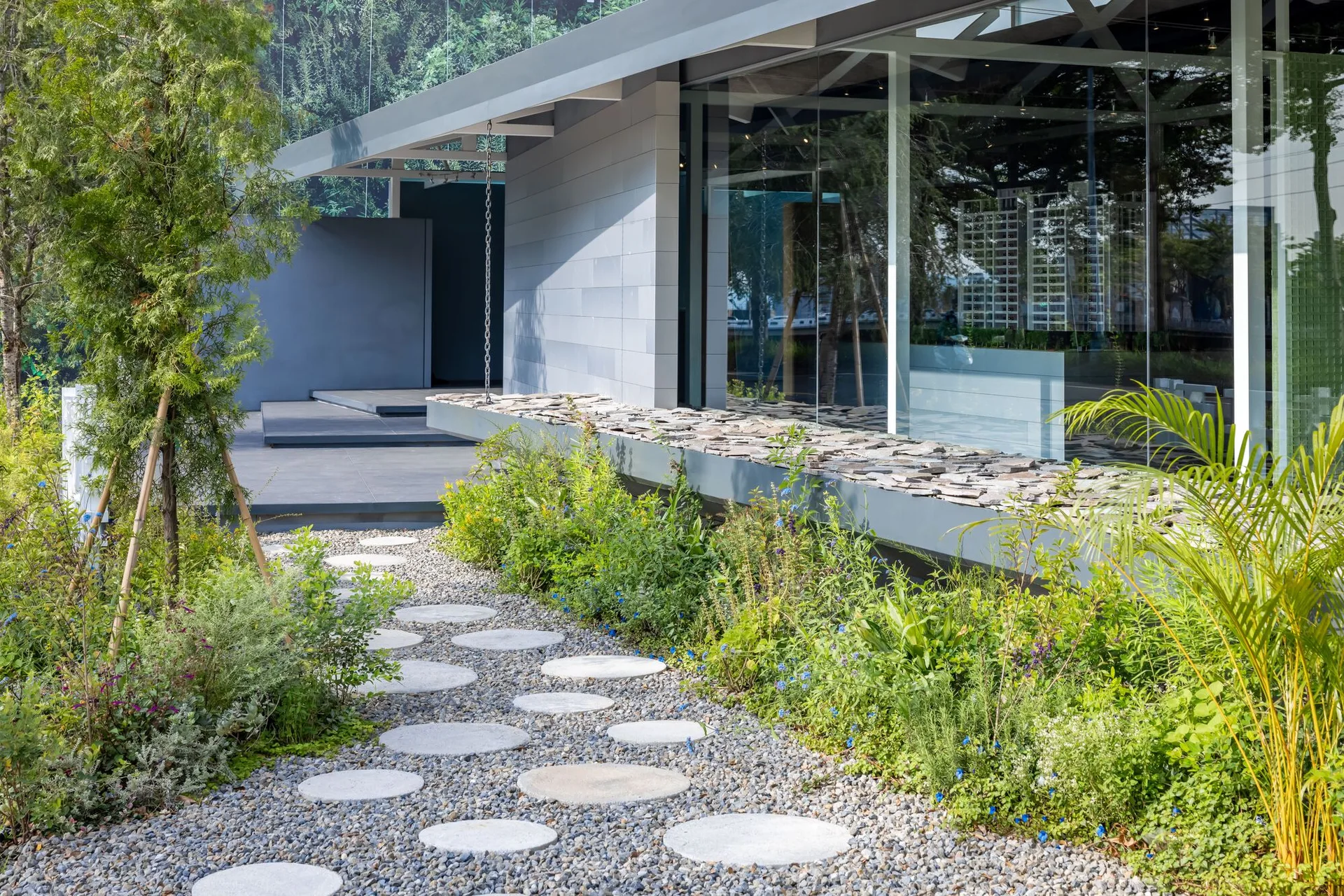 A modern grey building exterior features a gravel garden path with circular stone stepping stones alongside green plants and a large glass facade.