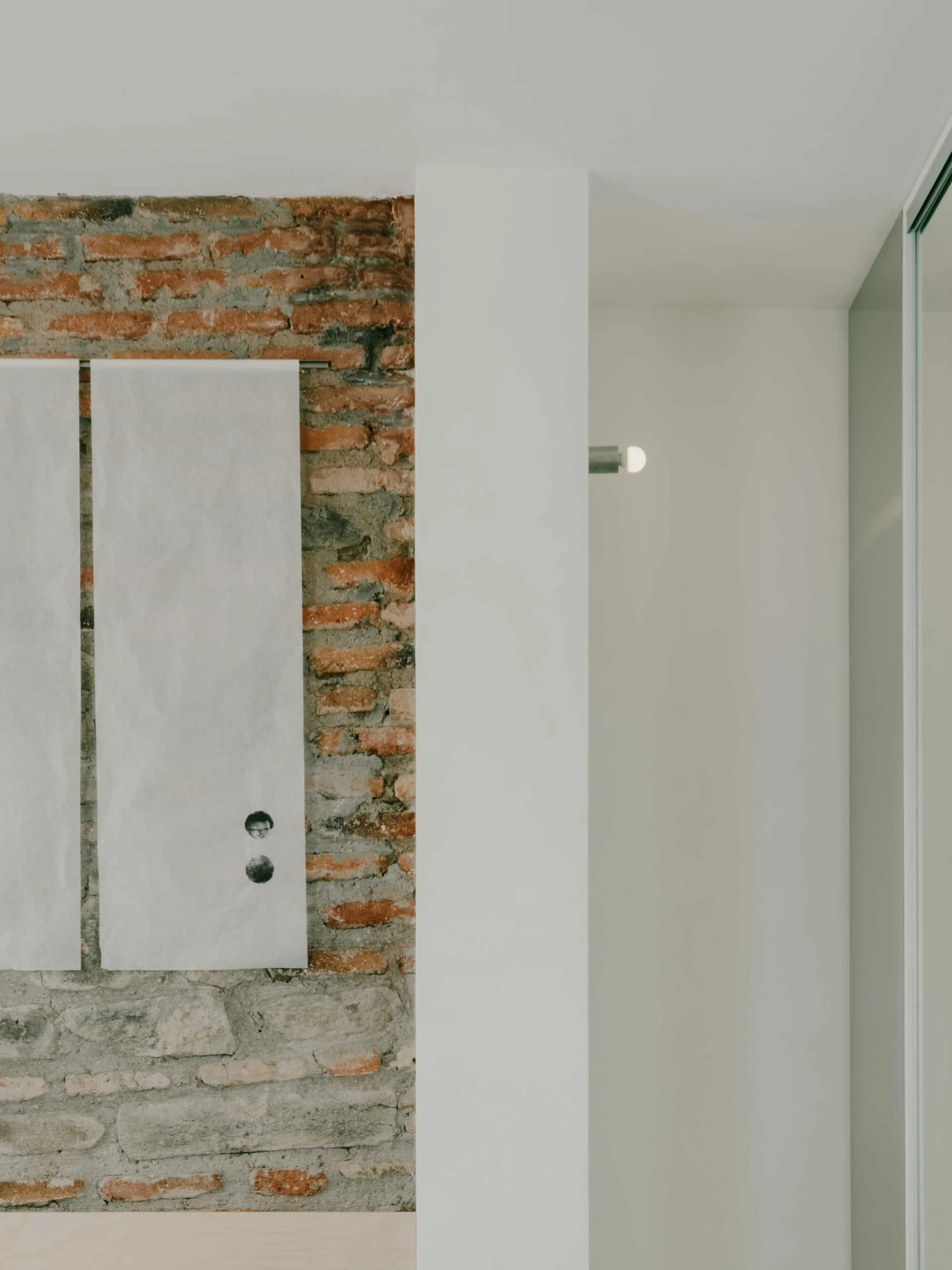 Two minimalist white paper banners hang on an exposed red brick wall next to a smooth white column with a small lit wall fixture.