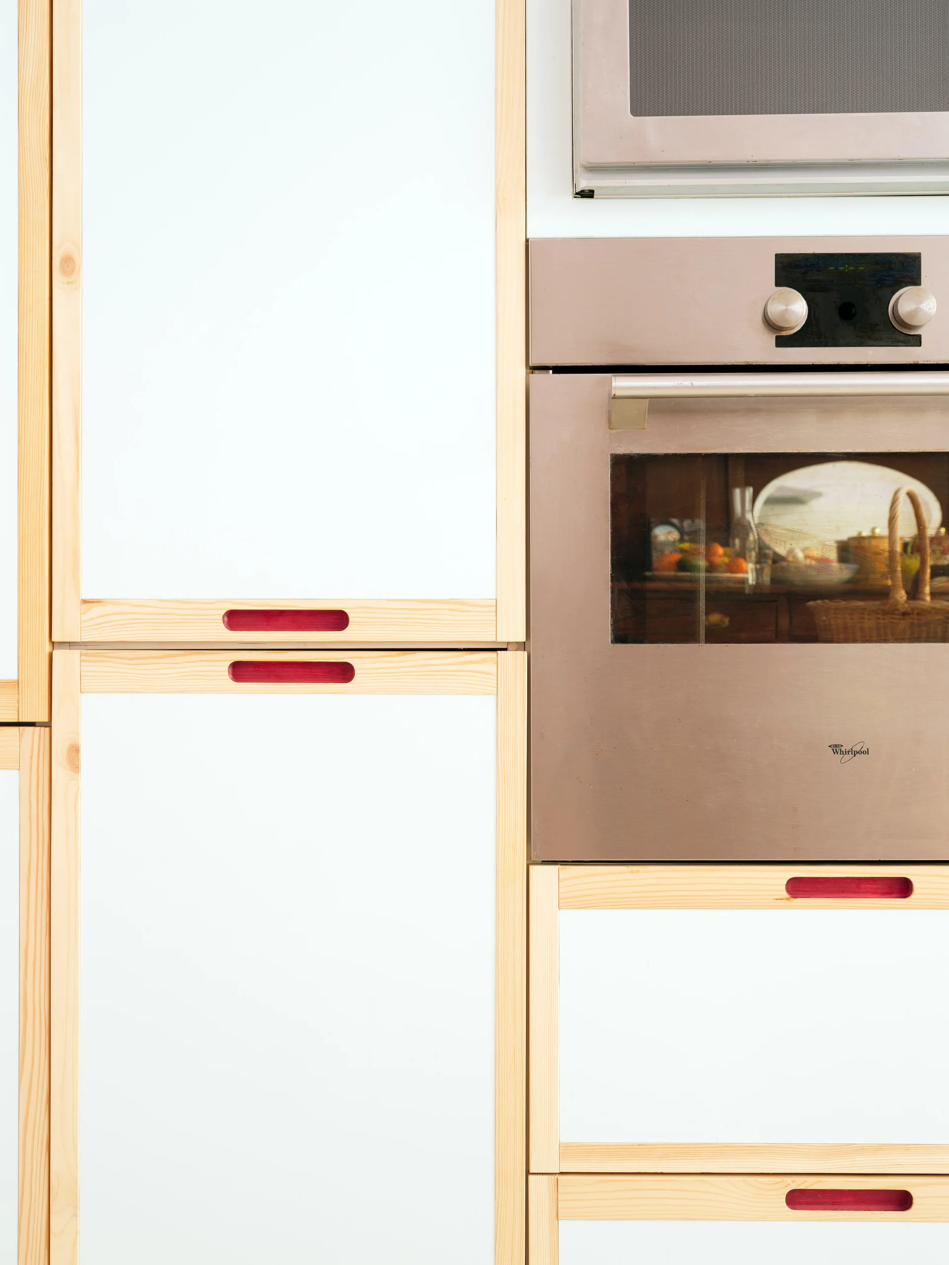 A close-up of white kitchen cabinet doors with light wood frames and red recessed handles next to a built-in stainless steel oven.