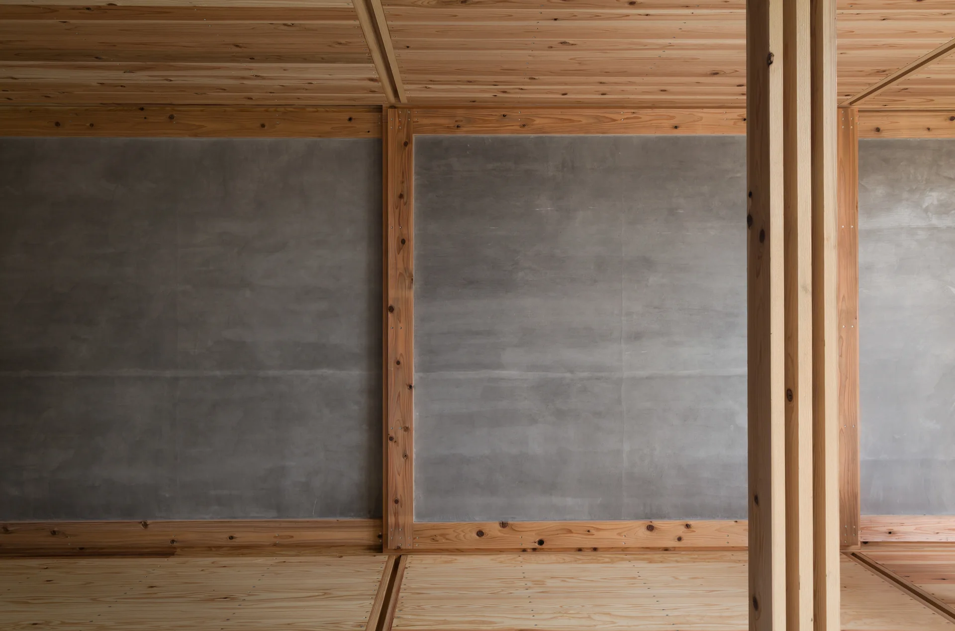 An unfurnished room features smooth gray concrete wall panels framed by light-toned exposed wood beams, columns, and matching wooden flooring and ceiling.